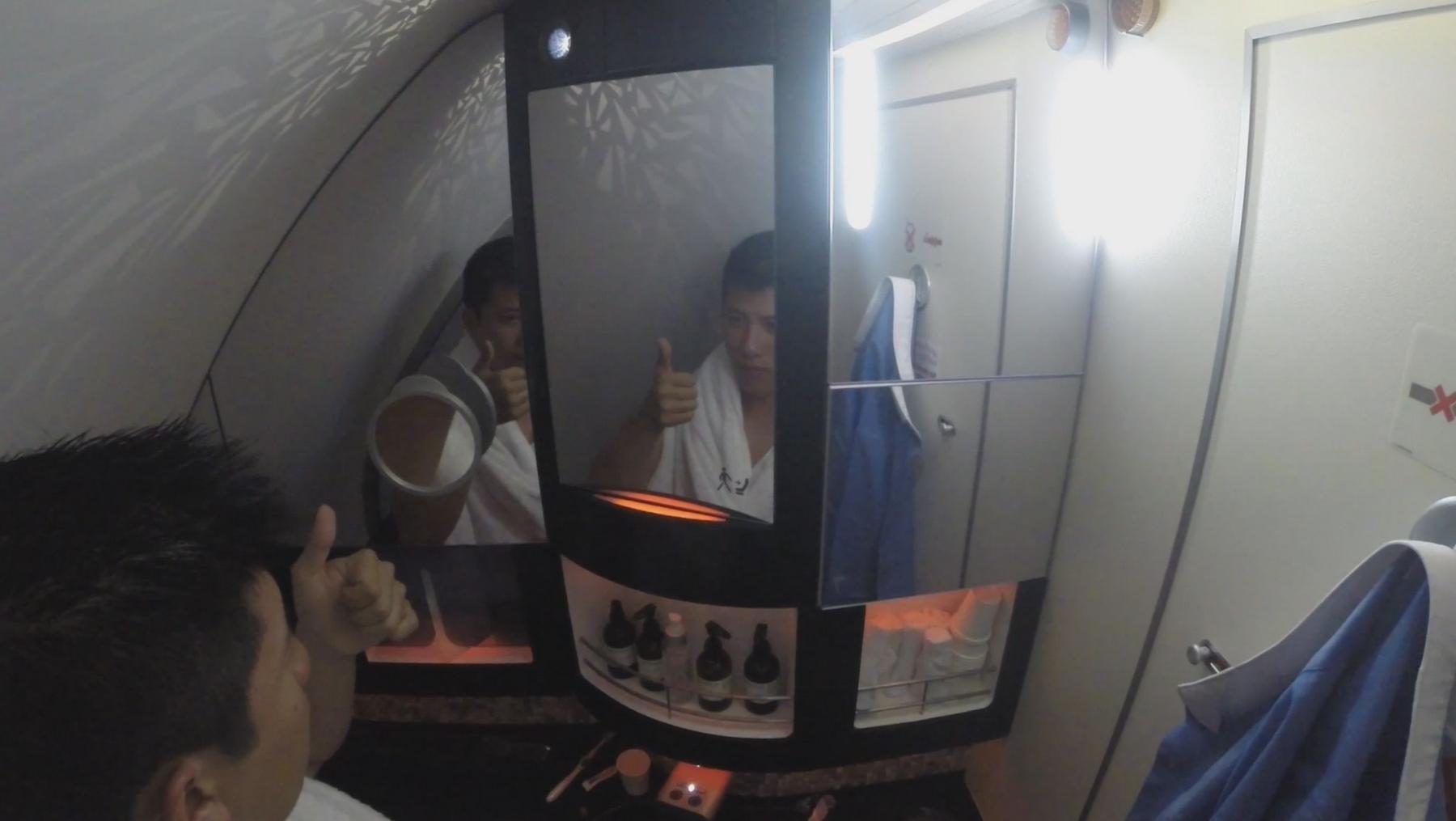 A man in a white towel gives a thumbs-up, reflected in a mirror inside an airplane vanity.