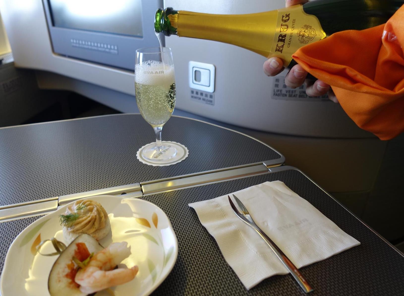A hand pours Krug champagne into a glass on an Eva Air business class tray table, with appetizers on a plate.