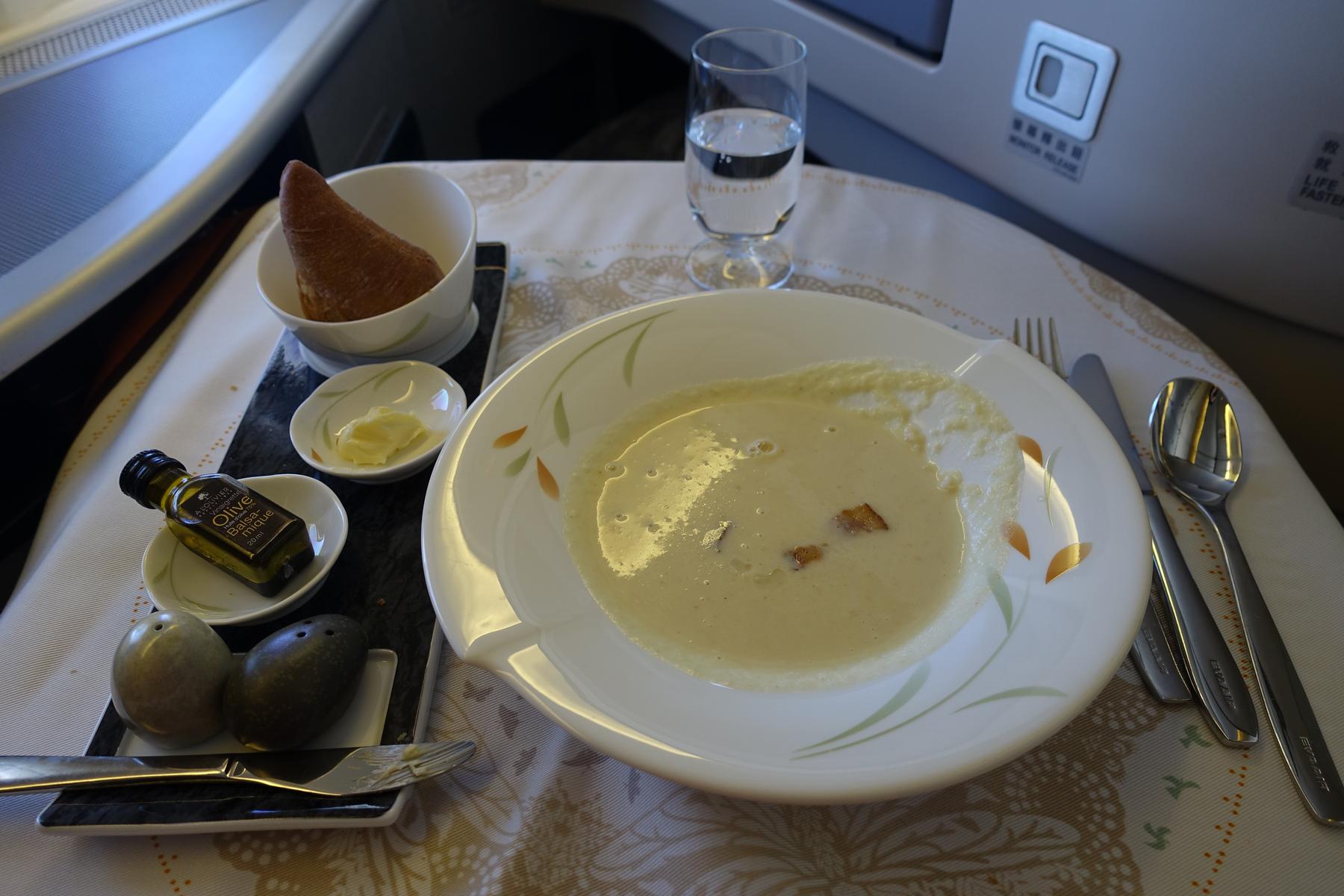 A bowl of soup, bread, and condiments are served on an airplane tray table in business class.