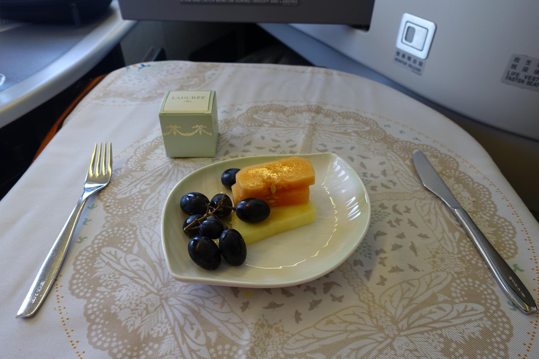 A plate with grapes, melon, and pineapple, a small Ladurée box, and cutlery sits on a patterned Eva Air Business Class tray table.