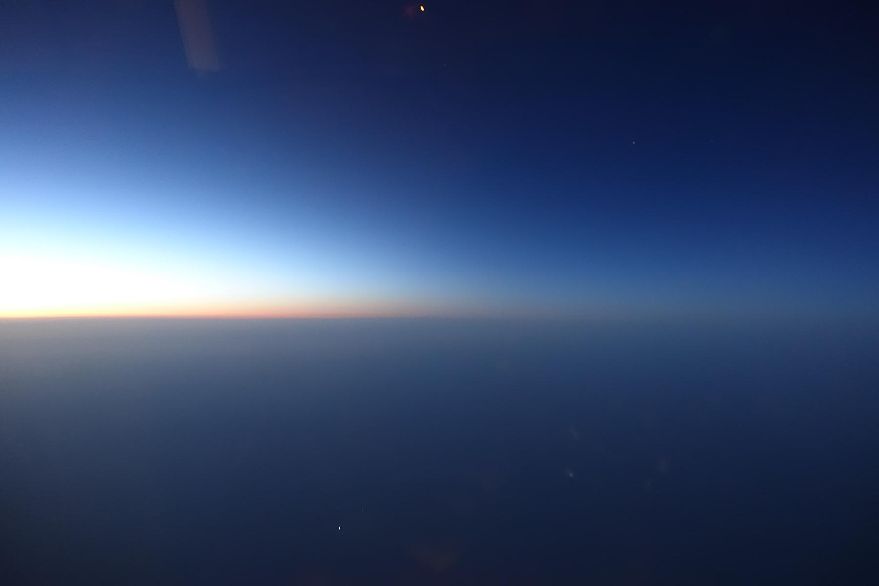 Airplane window view of the horizon at twilight, showing a gradient sky from pale orange to deep blue.