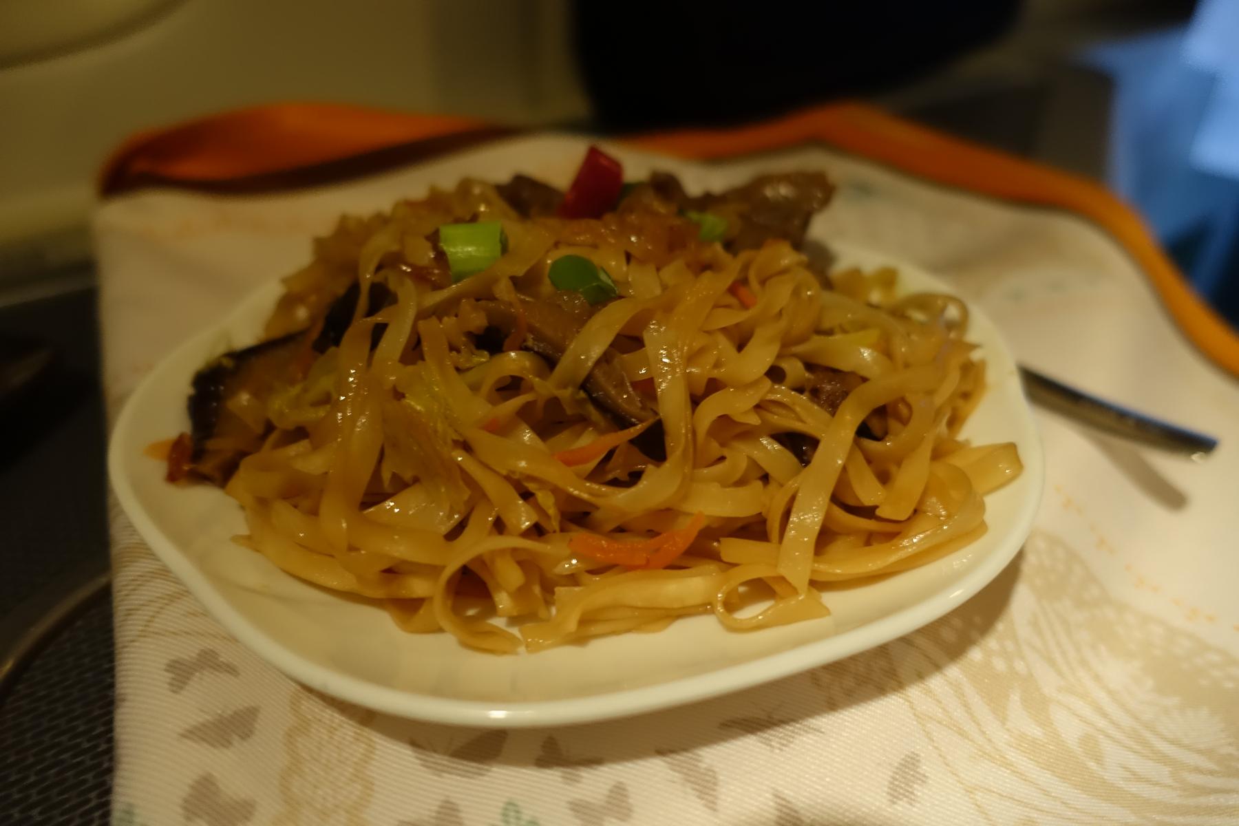 A white plate holds stir-fried flat noodles garnished with green onions and red pepper.
