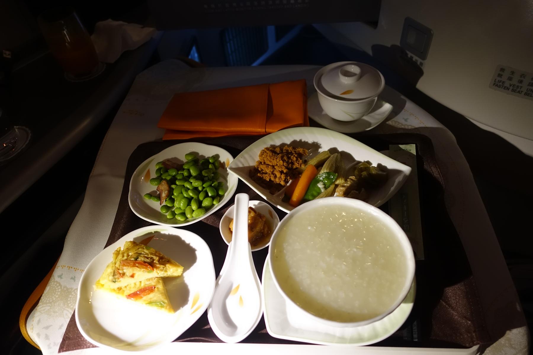 An Eva Air Business Class meal tray with several dishes, including porridge, omelet, edamame, and vegetables, in a dimly lit airplane cabin.