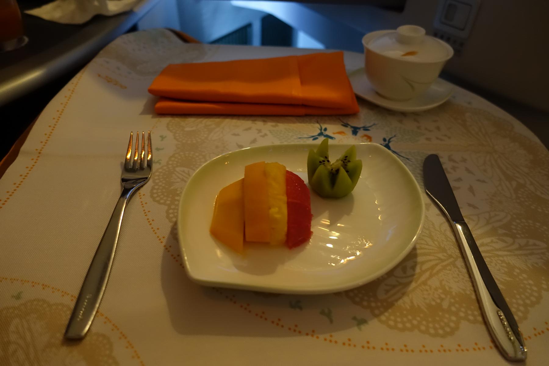 A plate of cut fruit, including carved kiwi, with cutlery, an orange napkin, and a lidded cup on an airplane tray table.