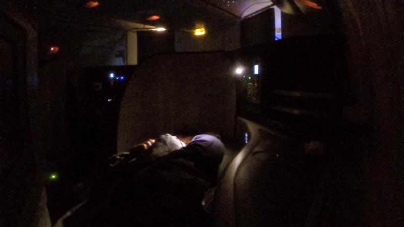 A passenger sleeps in a lie-flat business class seat in a dimly lit airplane cabin.