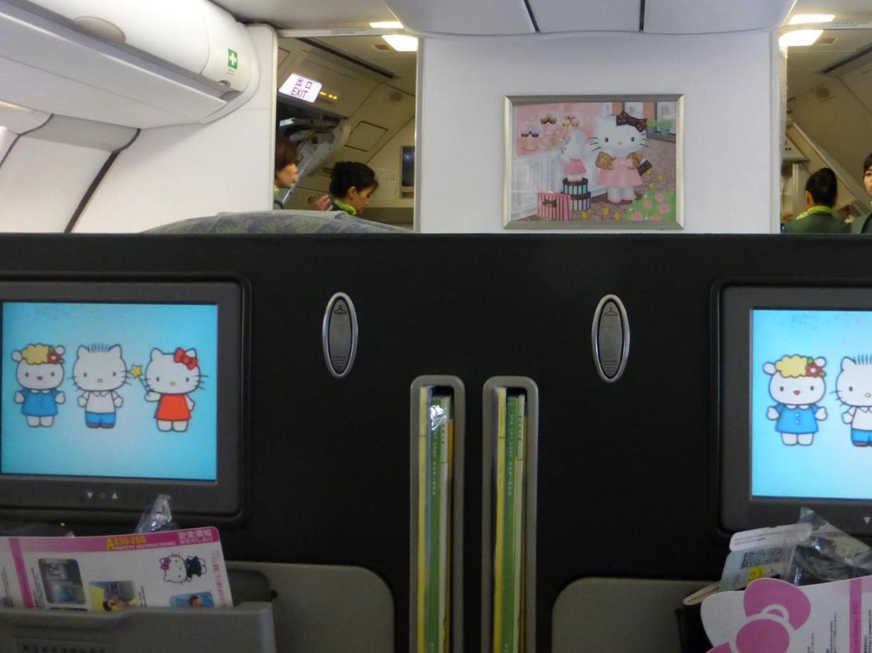 Airplane cabin interior featuring Hello Kitty themed entertainment screens and a framed picture, with flight attendants in the background.