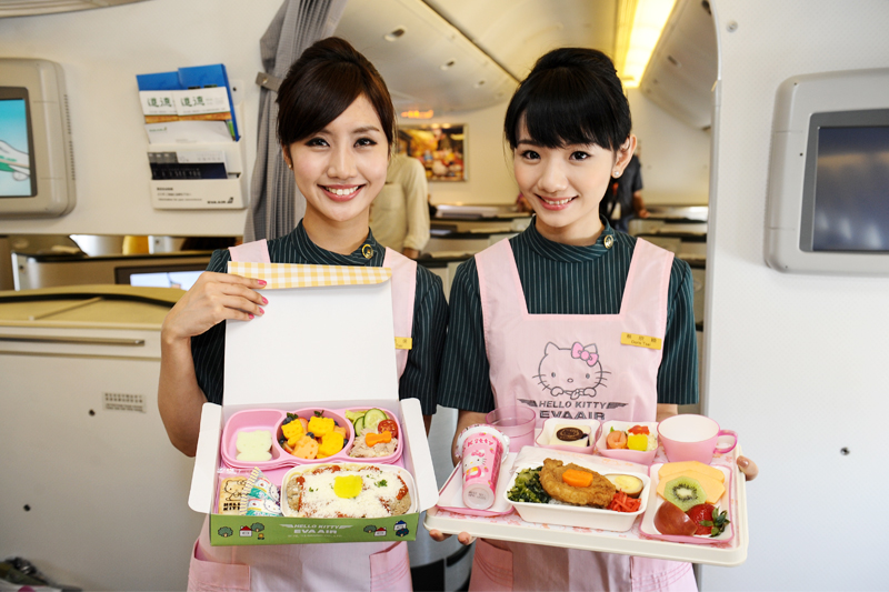 Two smiling flight attendants in Hello Kitty aprons present themed meals on an airplane.