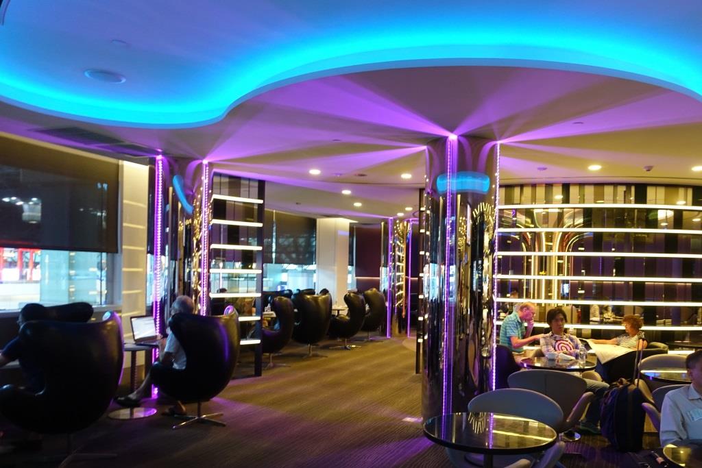 A stylish airport lounge with blue and purple ambient lighting and people relaxing.