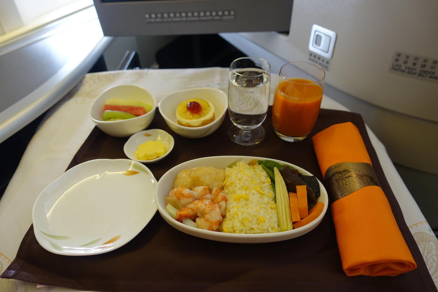 Eva Air business class meal on an airplane tray, with shrimp, rice, vegetables, fruit, dessert, and drinks.