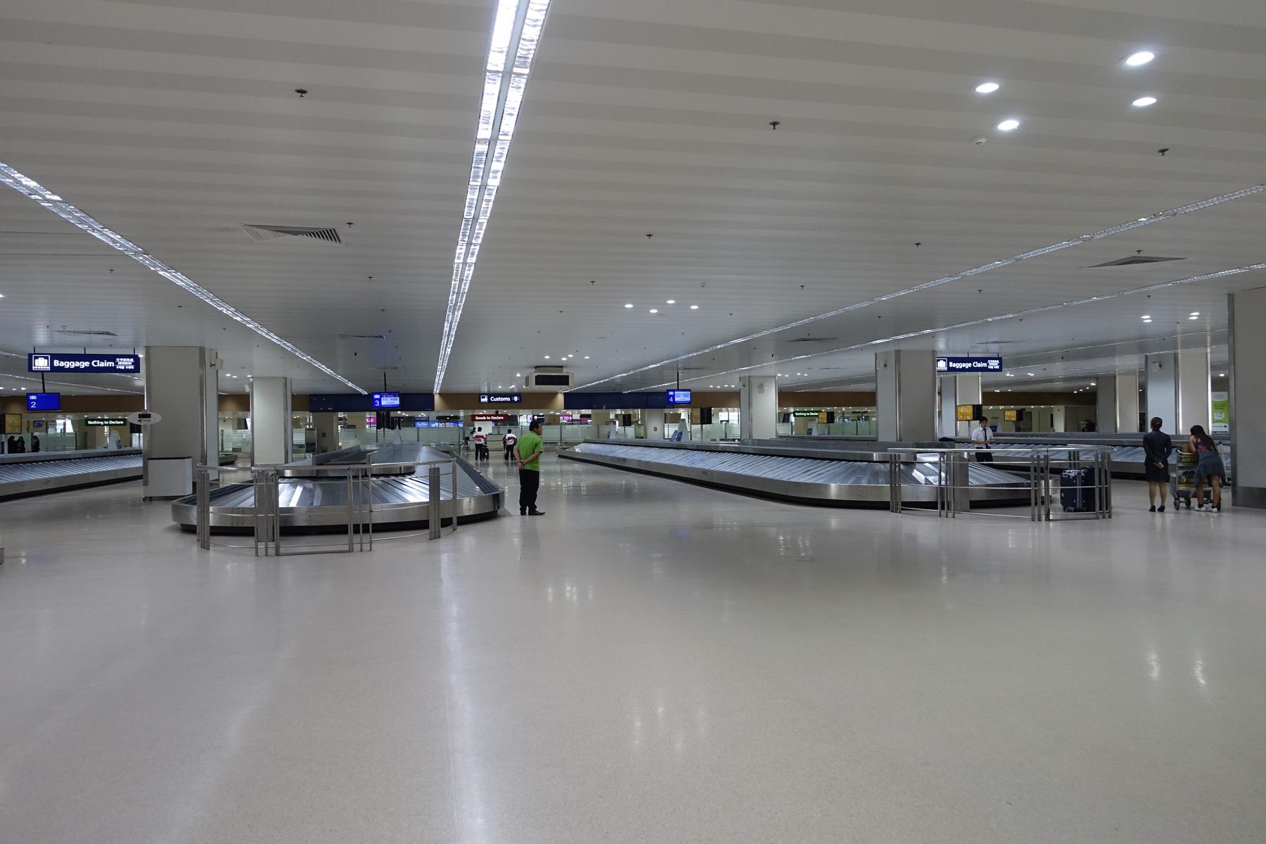 Airport baggage claim area with multiple carousels, overhead signs, and a few people.