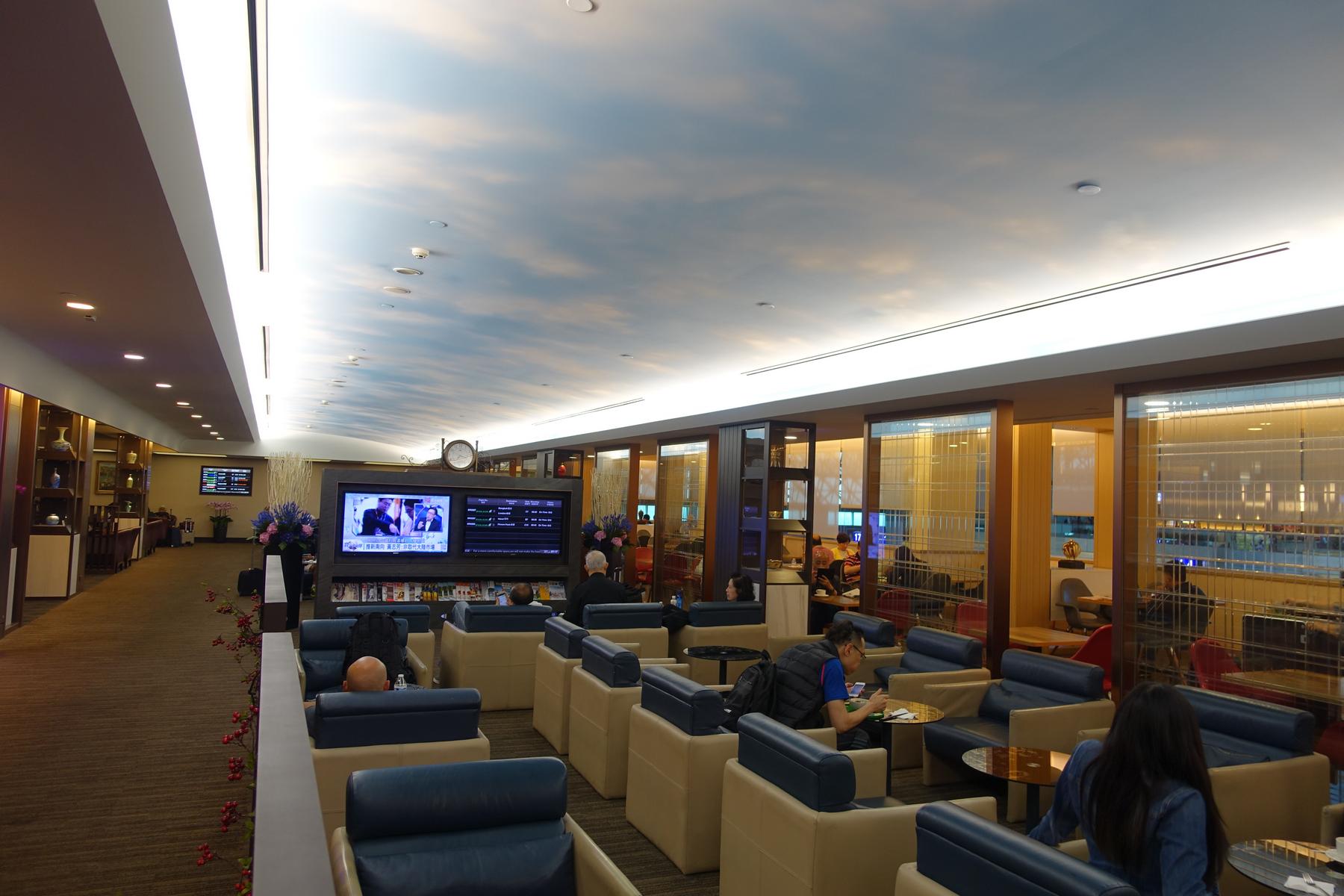 A spacious, modern business class lounge with a cloudy sky ceiling, multiple TV screens, and passengers relaxing in chairs.