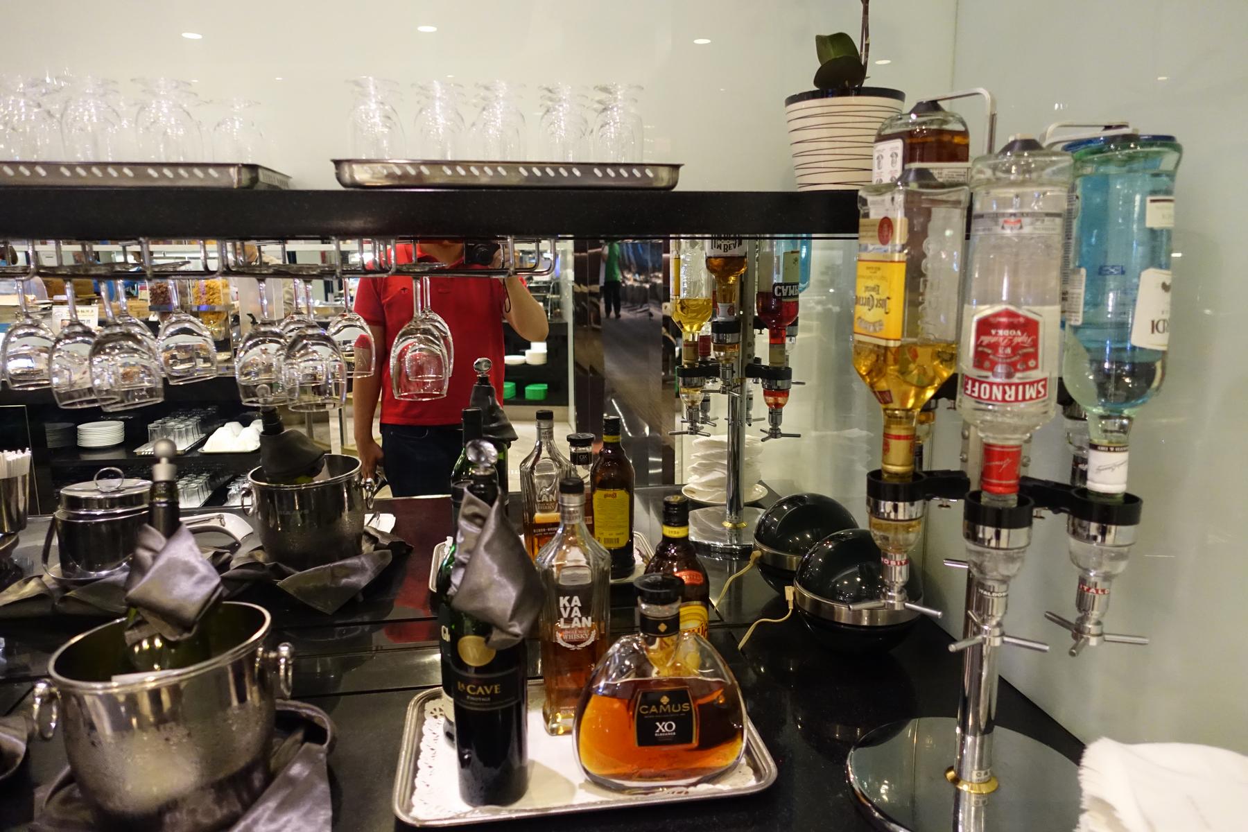 An airline business class lounge bar with liquor dispensers, bottled spirits, and hanging wine glasses.