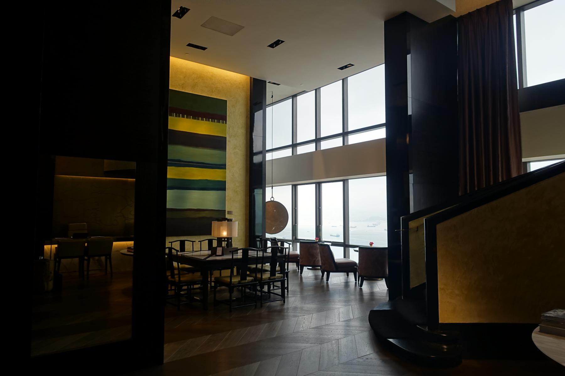 A modern, spacious hotel suite features a dining area, lounge seating, an abstract painting, and large windows with a harbor view.
