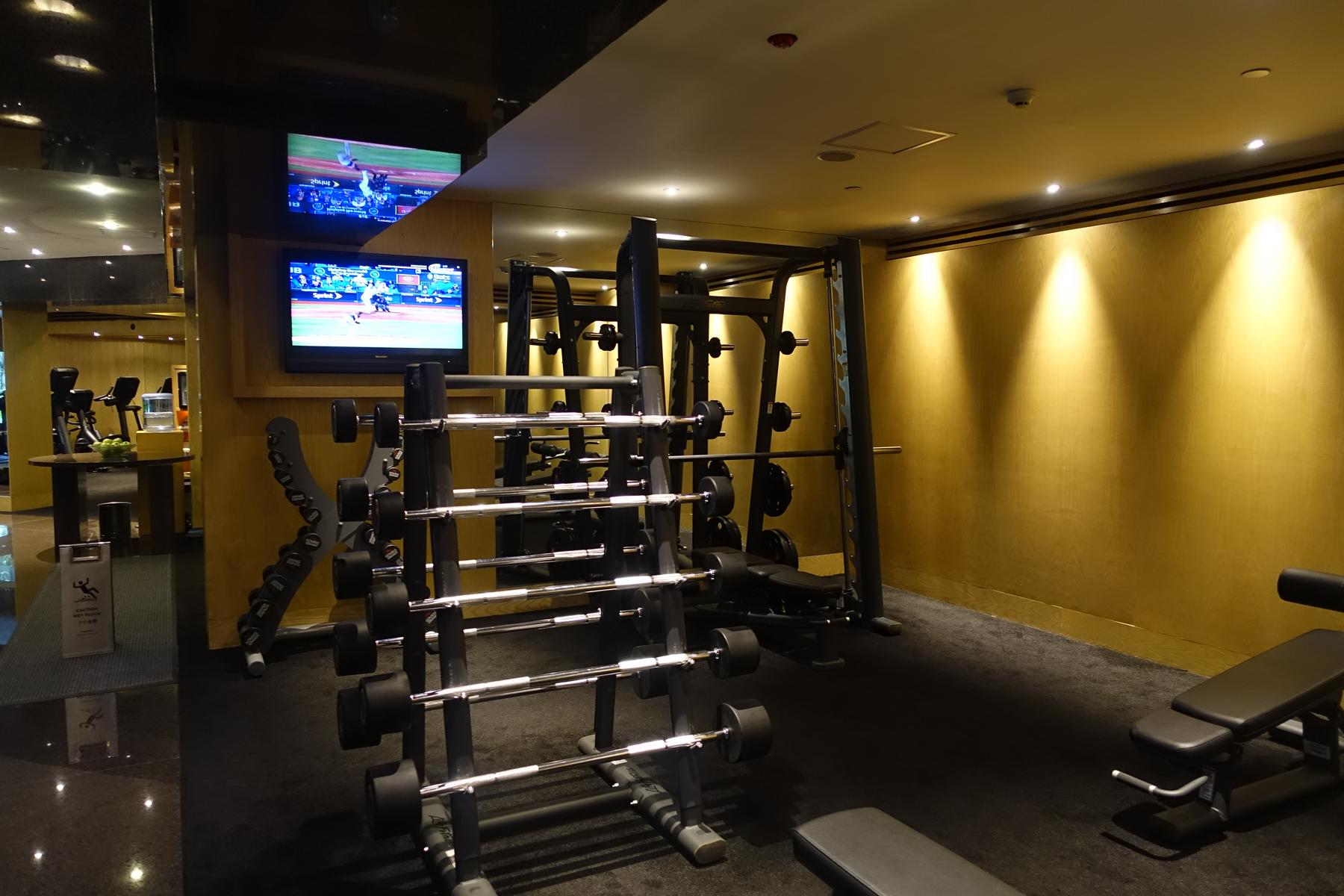A hotel gym with weight racks, exercise machines, and two televisions displaying a baseball game.