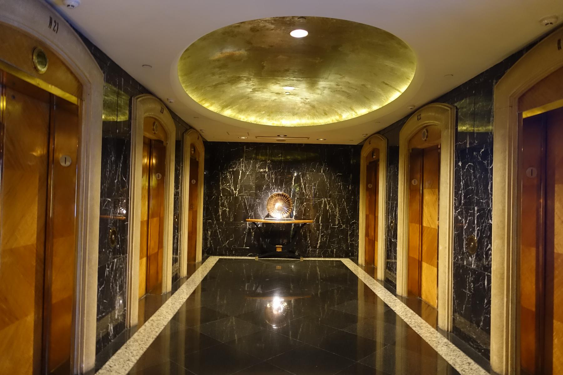 A lavish hotel elevator lobby with dark marble walls and floors, golden elevator doors, and a large illuminated gold dome on the ceiling.