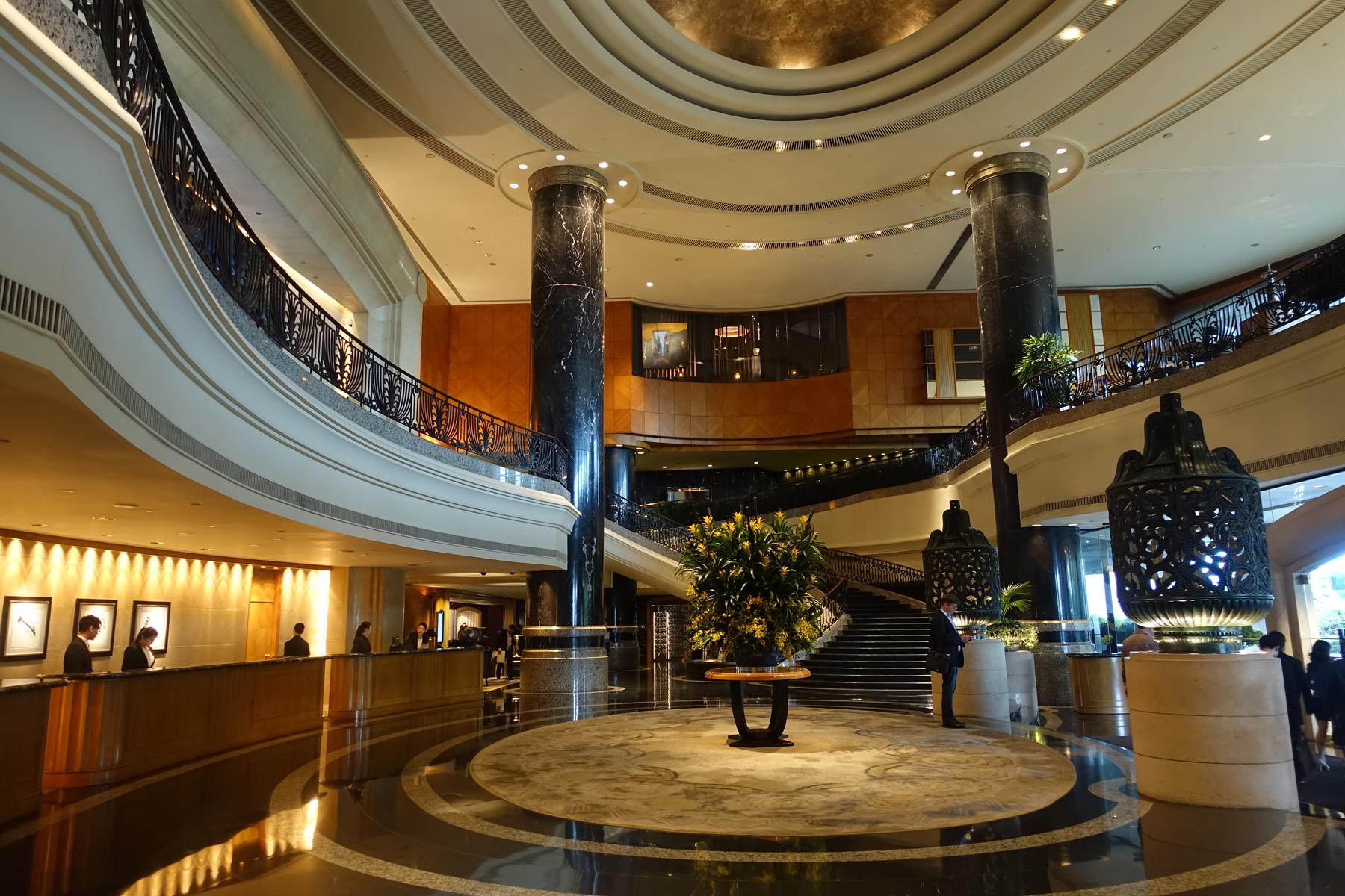 The Grand Hyatt Hong Kong's expansive, multi-story lobby features elegant staircases, large columns, and a high, ornate ceiling.