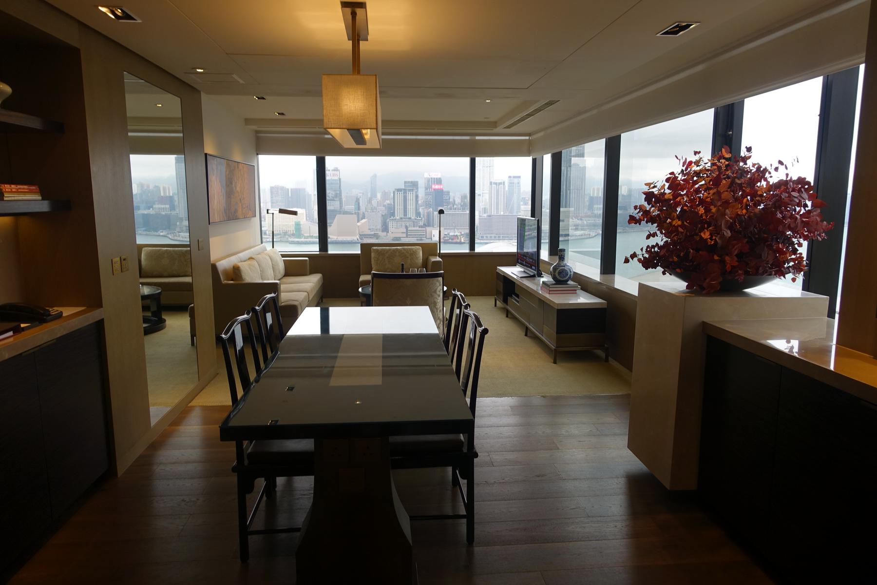 A spacious hotel suite featuring a dining area, living room, and expansive windows offering a city and harbor view.