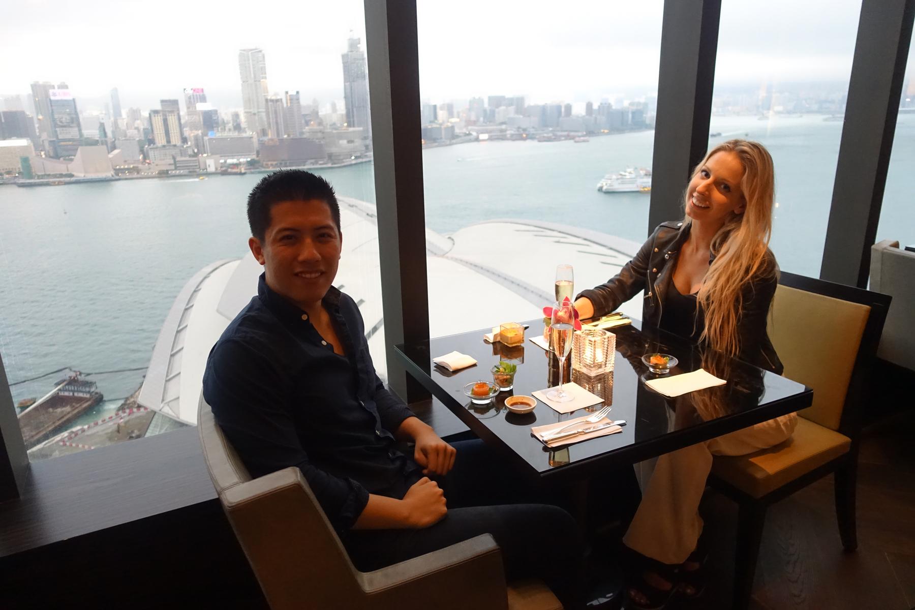 A couple sits at a table in the Grand Club Lounge with a panoramic view of Hong Kong's city and harbor.