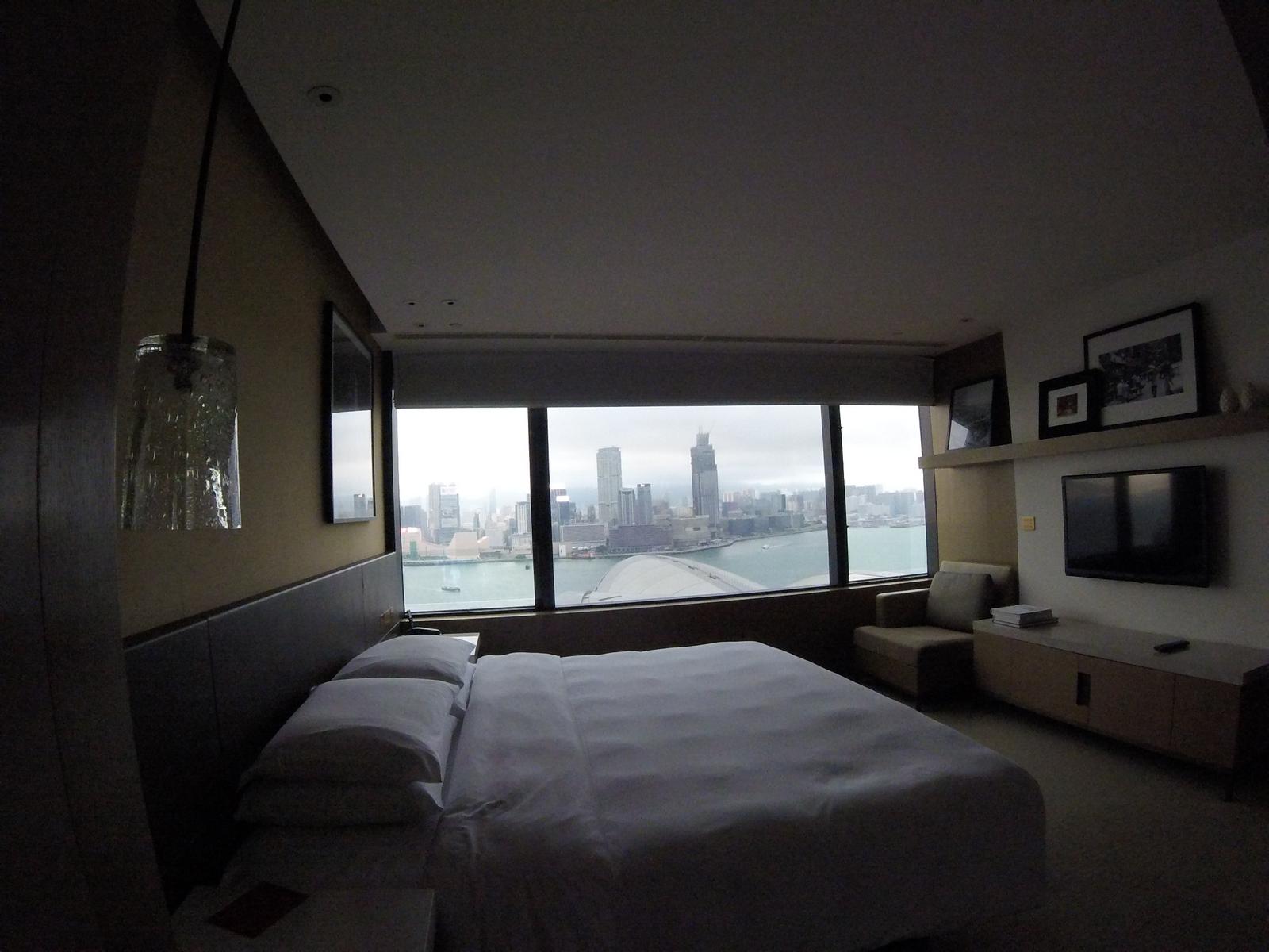 A hotel room with a large bed, a TV, and floor-to-ceiling windows overlooking the Hong Kong skyline and Victoria Harbour.