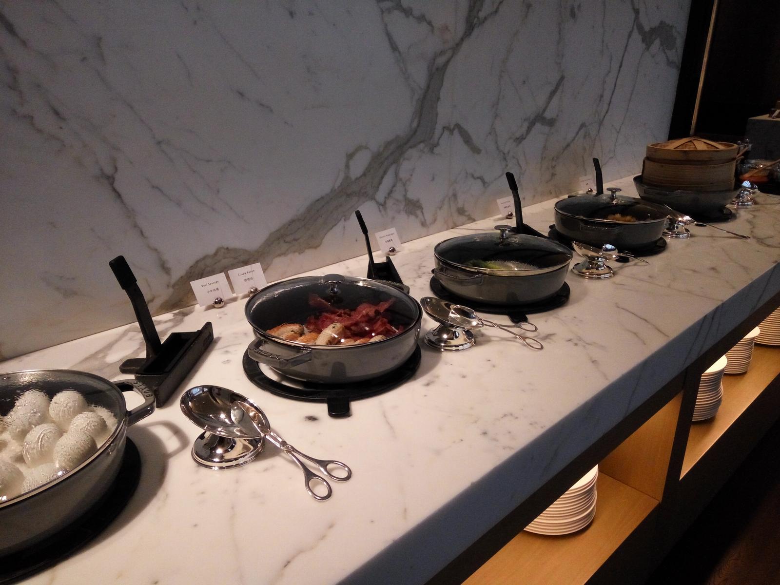 A buffet spread featuring hot dishes like bacon, sausages, and dim sum in covered pots on a white marble counter.