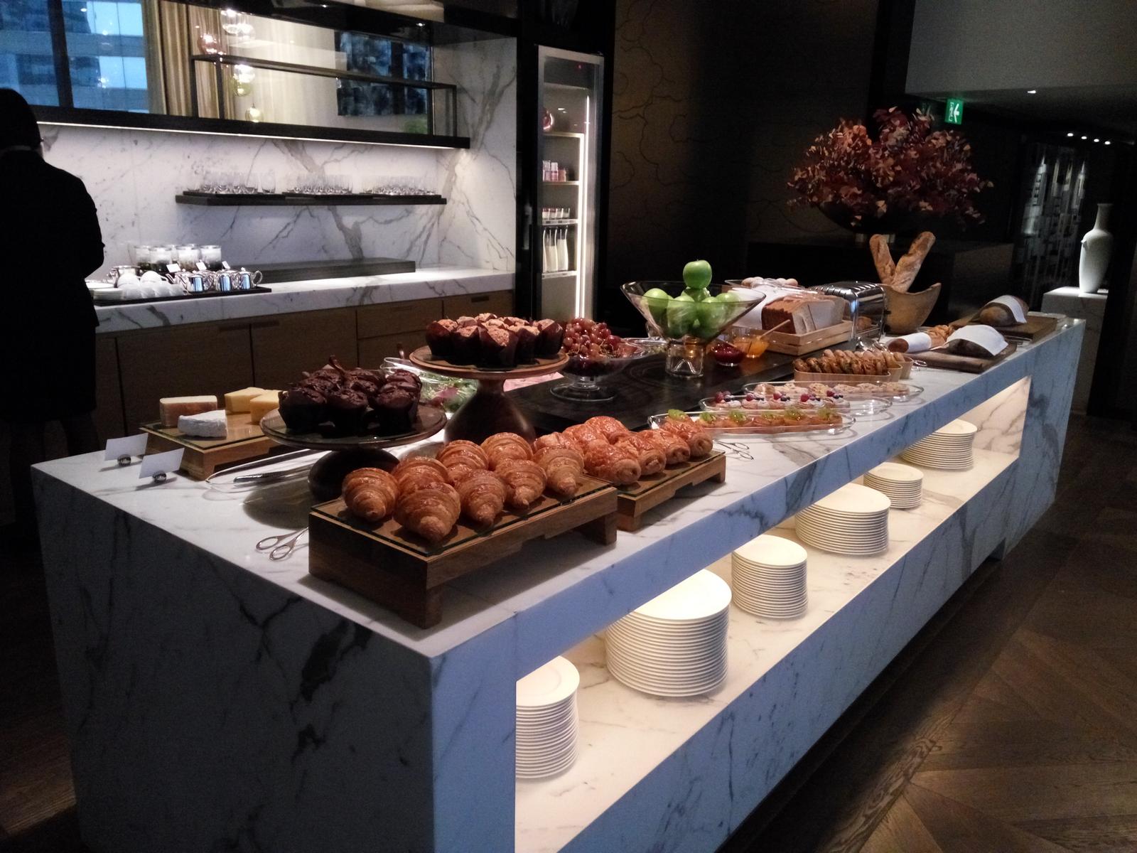 A hotel lounge buffet with pastries, fruits, and cheese on a marble counter.