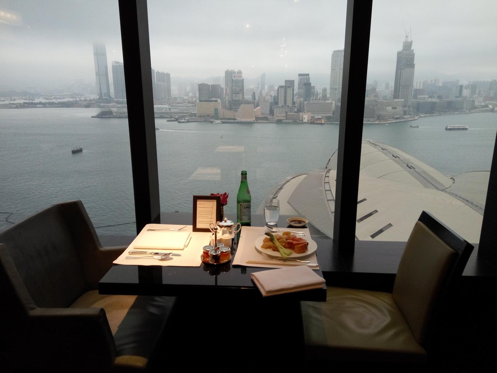 A dining table set with a meal and drinks faces a large window offering expansive views of a city harbor and skyline.