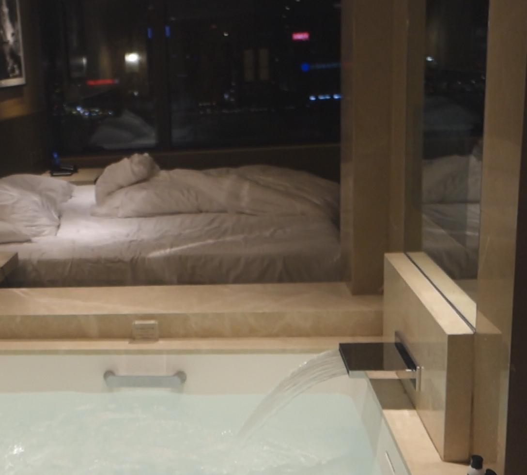 A luxurious bathtub with a waterfall faucet fills, overlooking a hotel bedroom and a city nightscape.