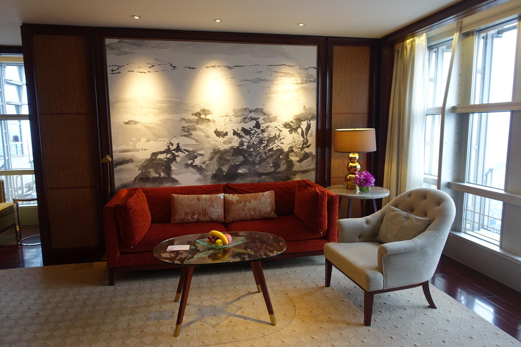 A stylish hotel living area features a red sofa, beige armchair, an oval coffee table with fruit, and a large abstract landscape wall mural.