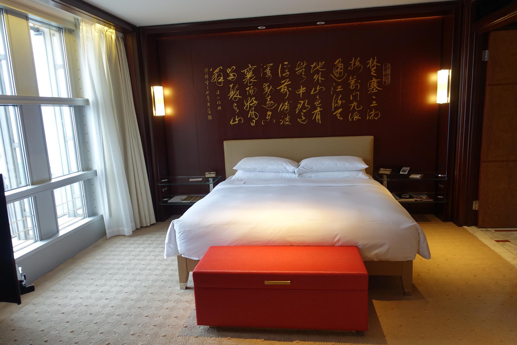A hotel room featuring a large bed, a red accent wall with golden Chinese calligraphy, and tall windows.