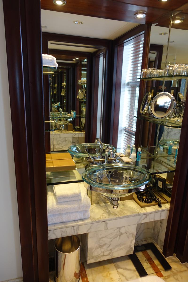 A hotel bathroom features dual clear glass sinks on a marble counter, reflecting in surrounding mirrors.
