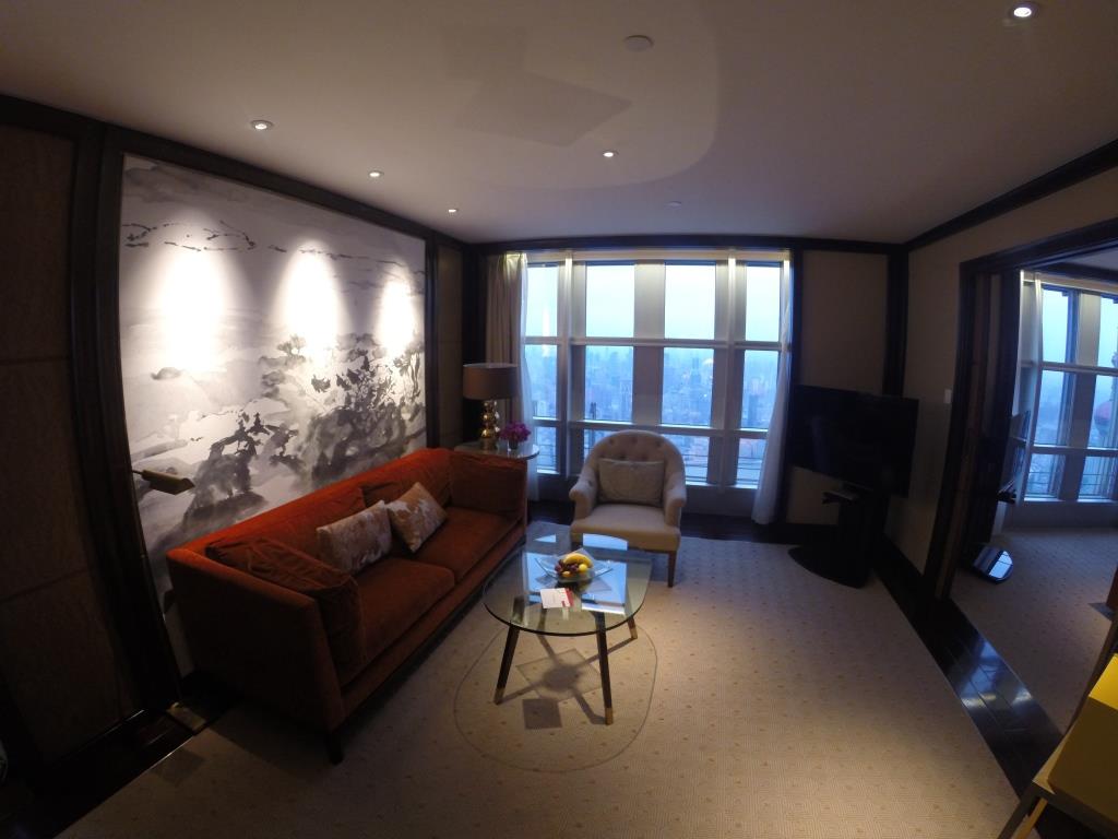 An elegant hotel suite living room featuring a red sofa, a large mural, and city views from wide windows.