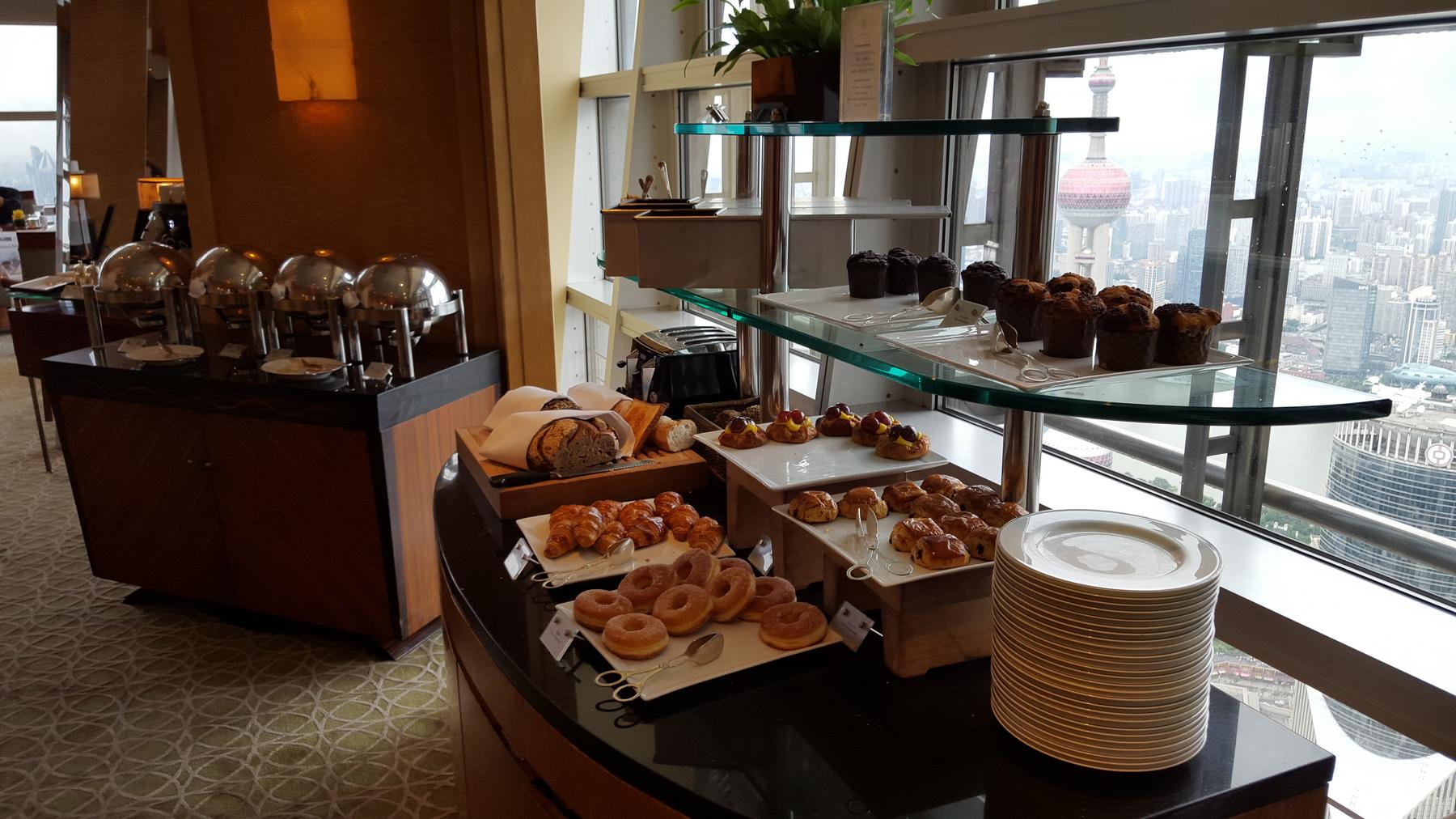 A breakfast buffet with pastries and hot food against a panoramic city view of Shanghai, including the Oriental Pearl Tower.