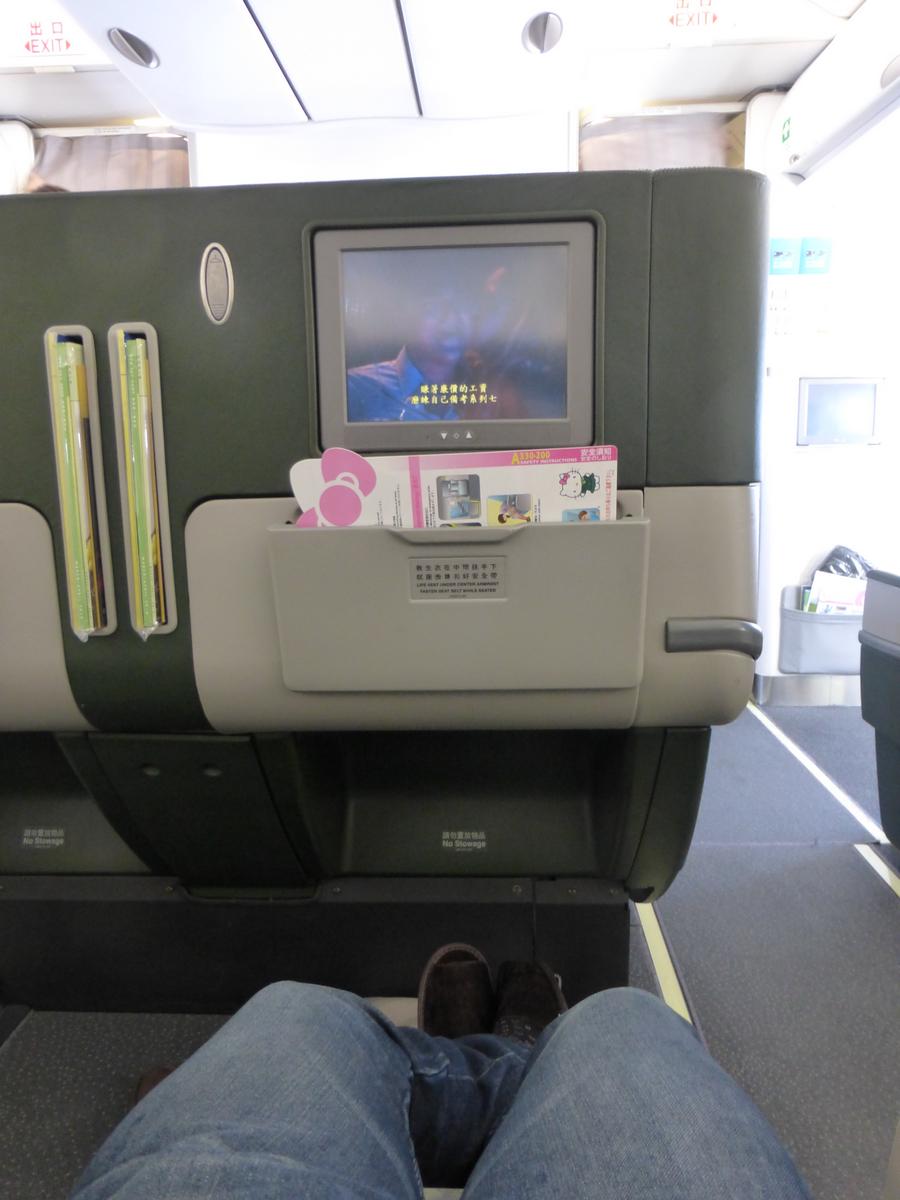 First-person view of a passenger's legs extended in a business class airplane seat, with an entertainment screen and Hello Kitty magazine.