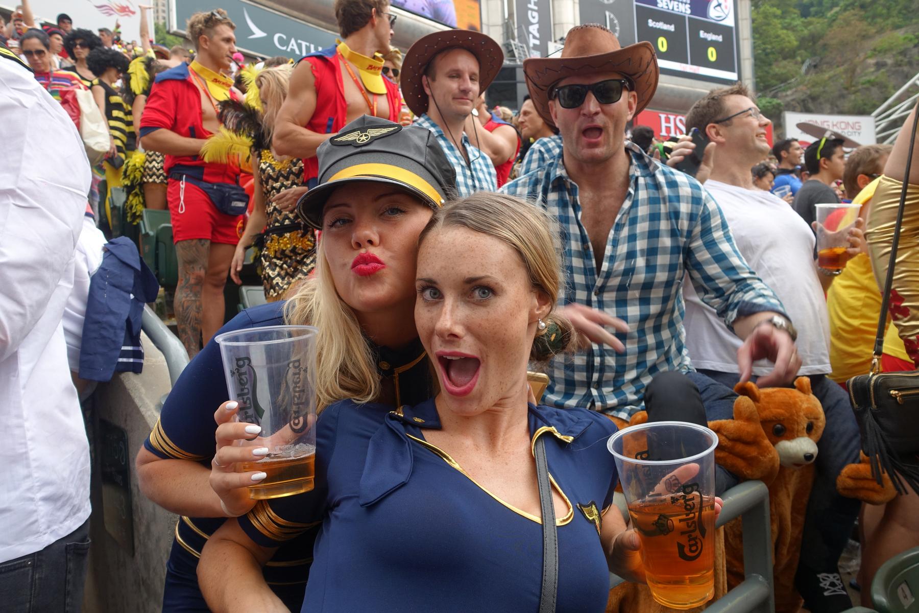 Two women in flight attendant costumes hold beers and make animated faces in the crowded South Stand at the Hong Kong Rugby Sevens.