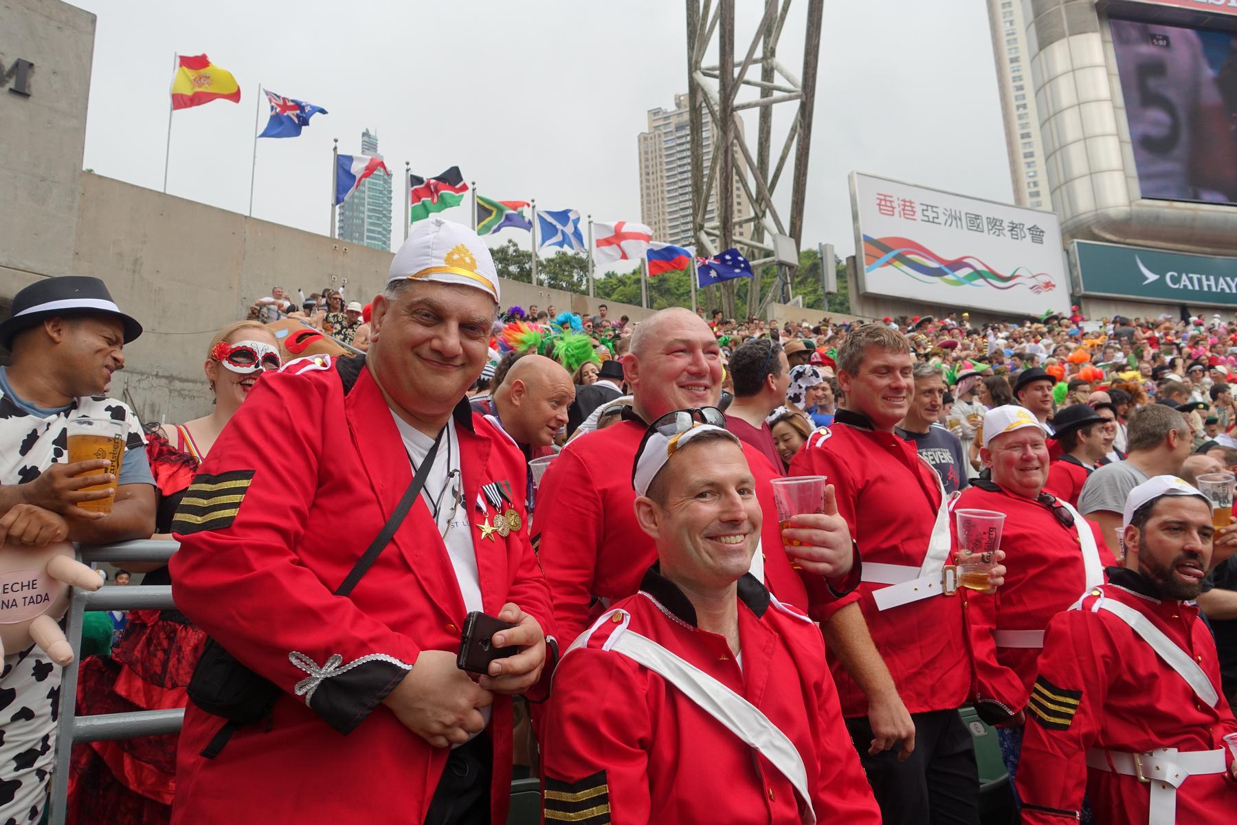 Cheerful spectators, including men in red uniform-like costumes, enjoy the Hong Kong Rugby Sevens.