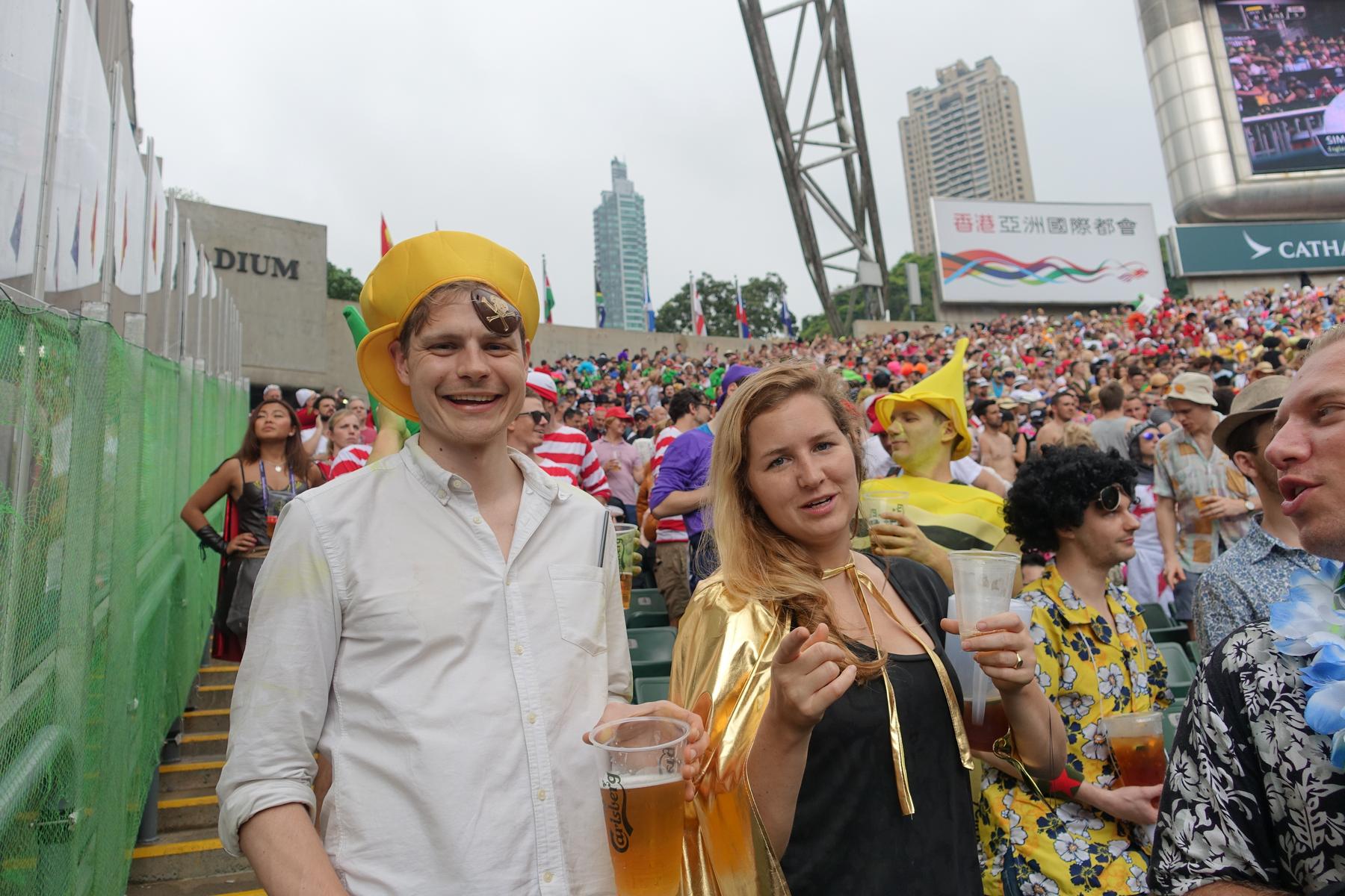 Two smiling spectators in costume, holding drinks, in the lively South Stand crowd at the Hong Kong Rugby Sevens.