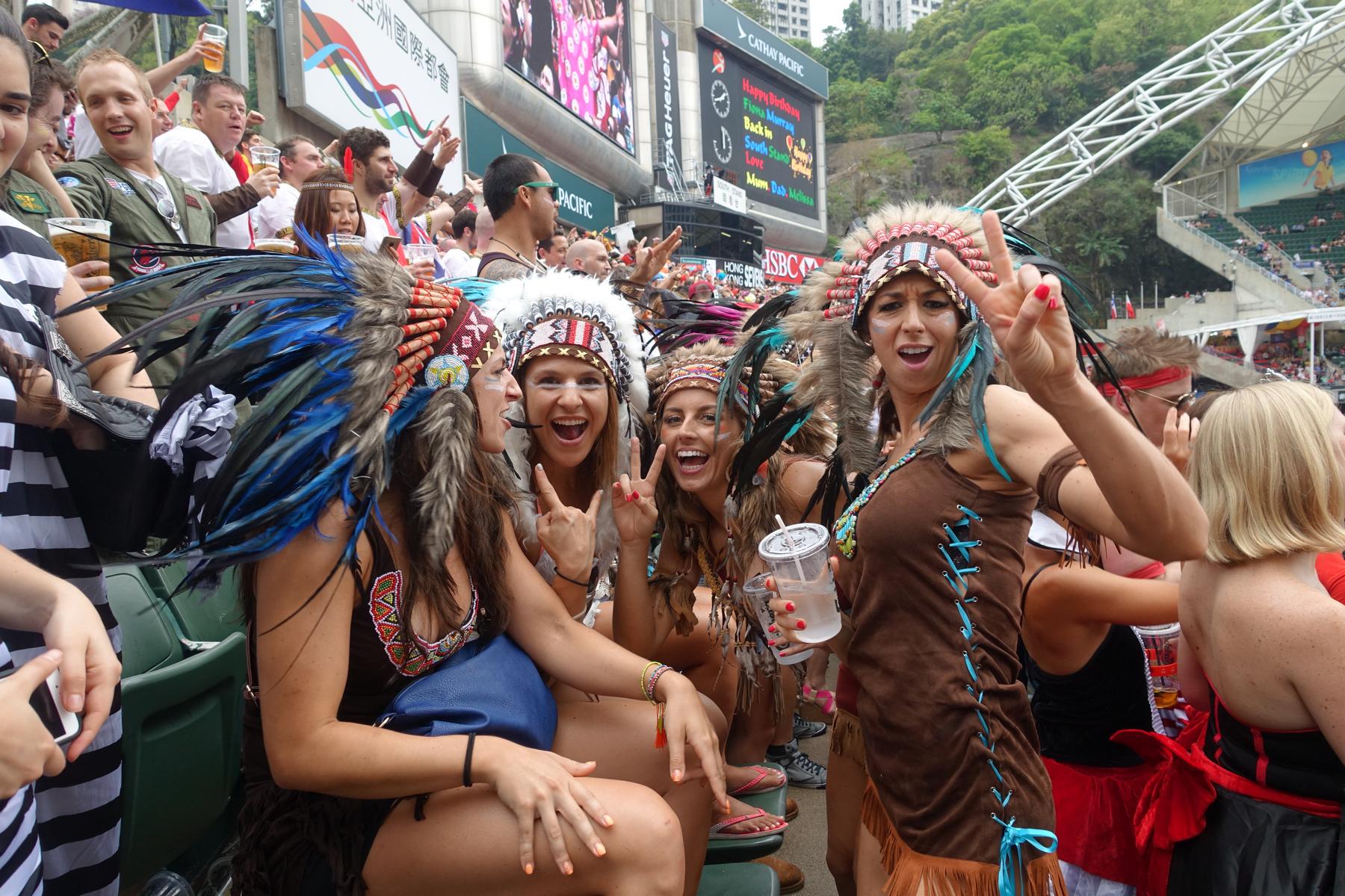 Four women in elaborate feathered headdresses smile and pose at the Hong Kong Rugby Sevens South Stand.