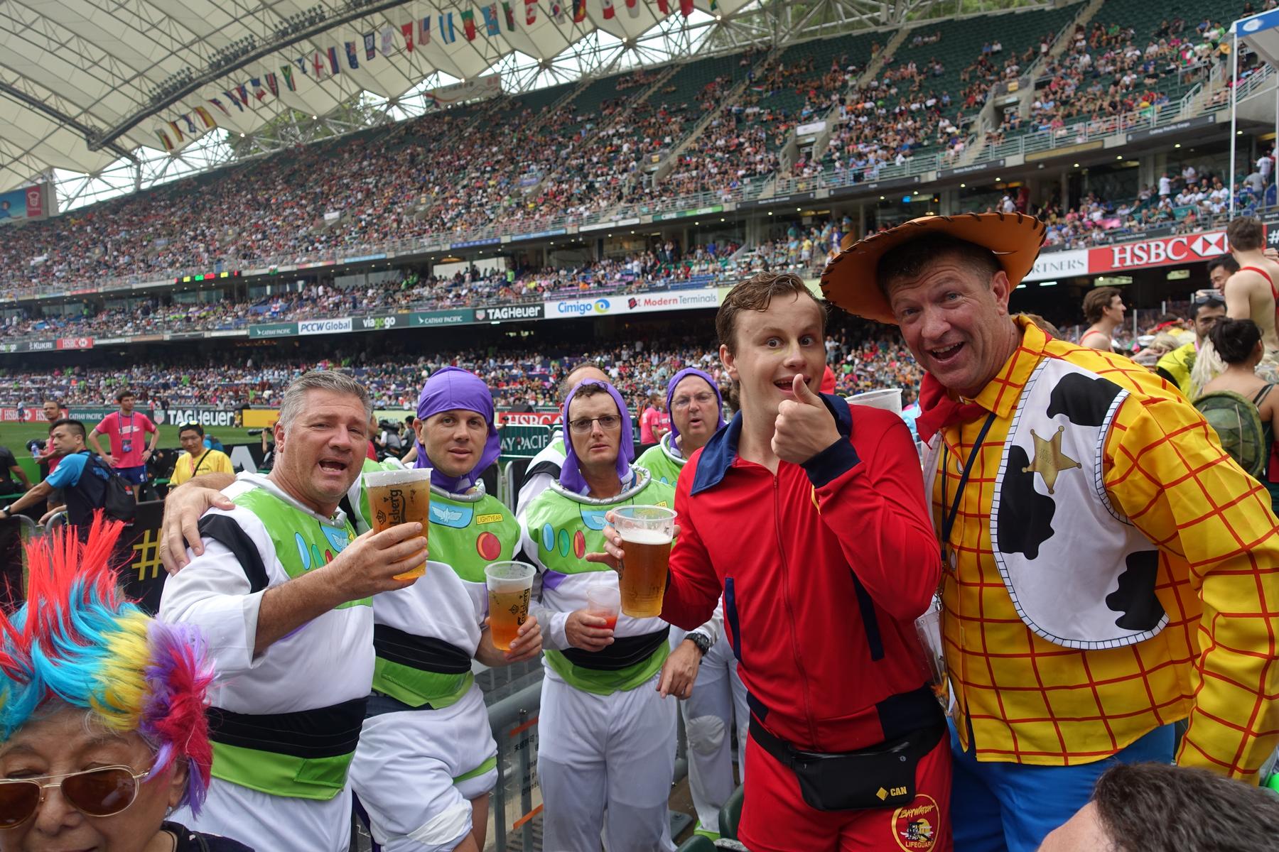 Costumed fans dressed as Woody and Buzz Lightyear hold drinks and pose in a crowded rugby stadium.