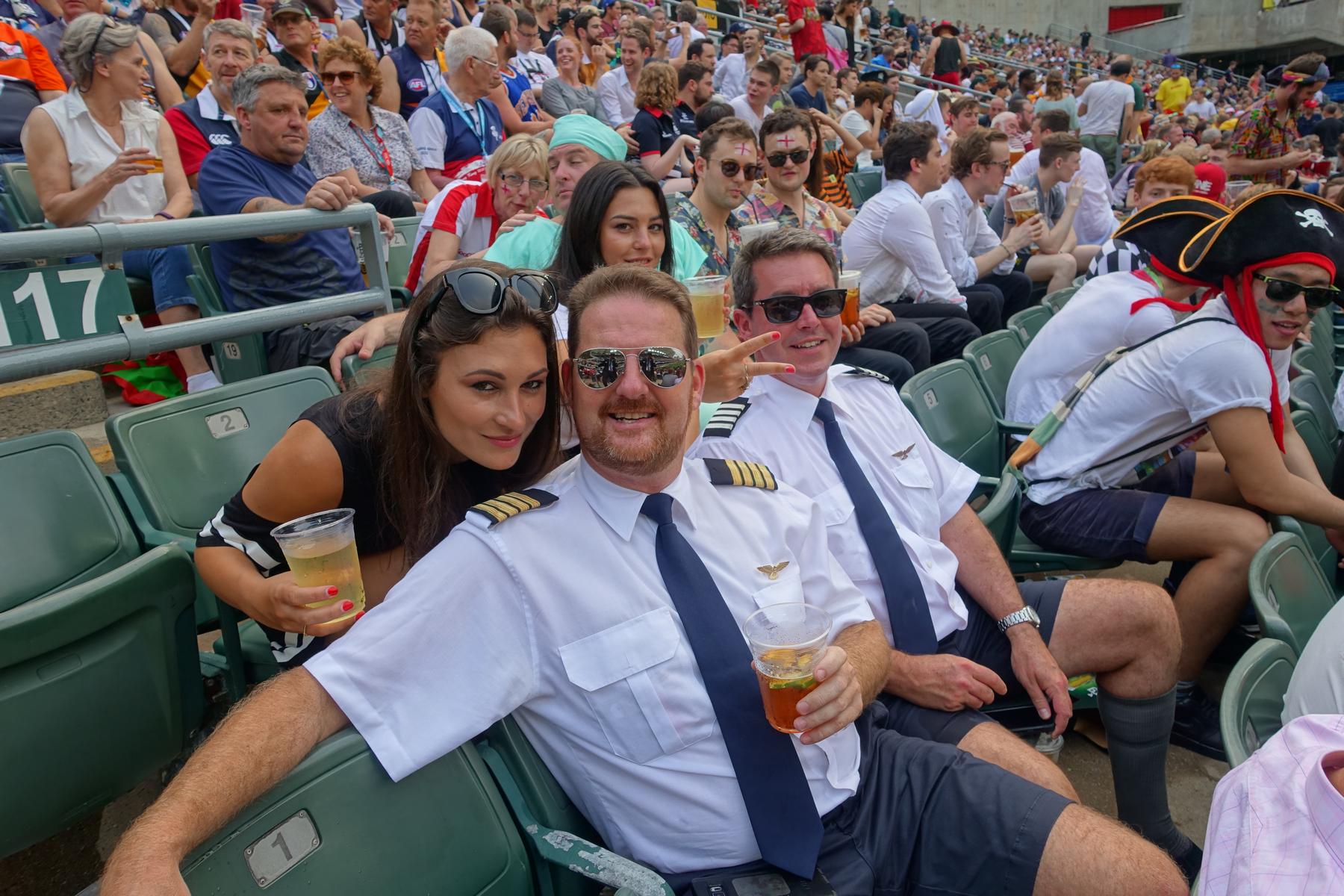 Fans in costumes, including two men dressed as pilots, smile and hold drinks in the festive South Stand at the Hong Kong Rugby Sevens.