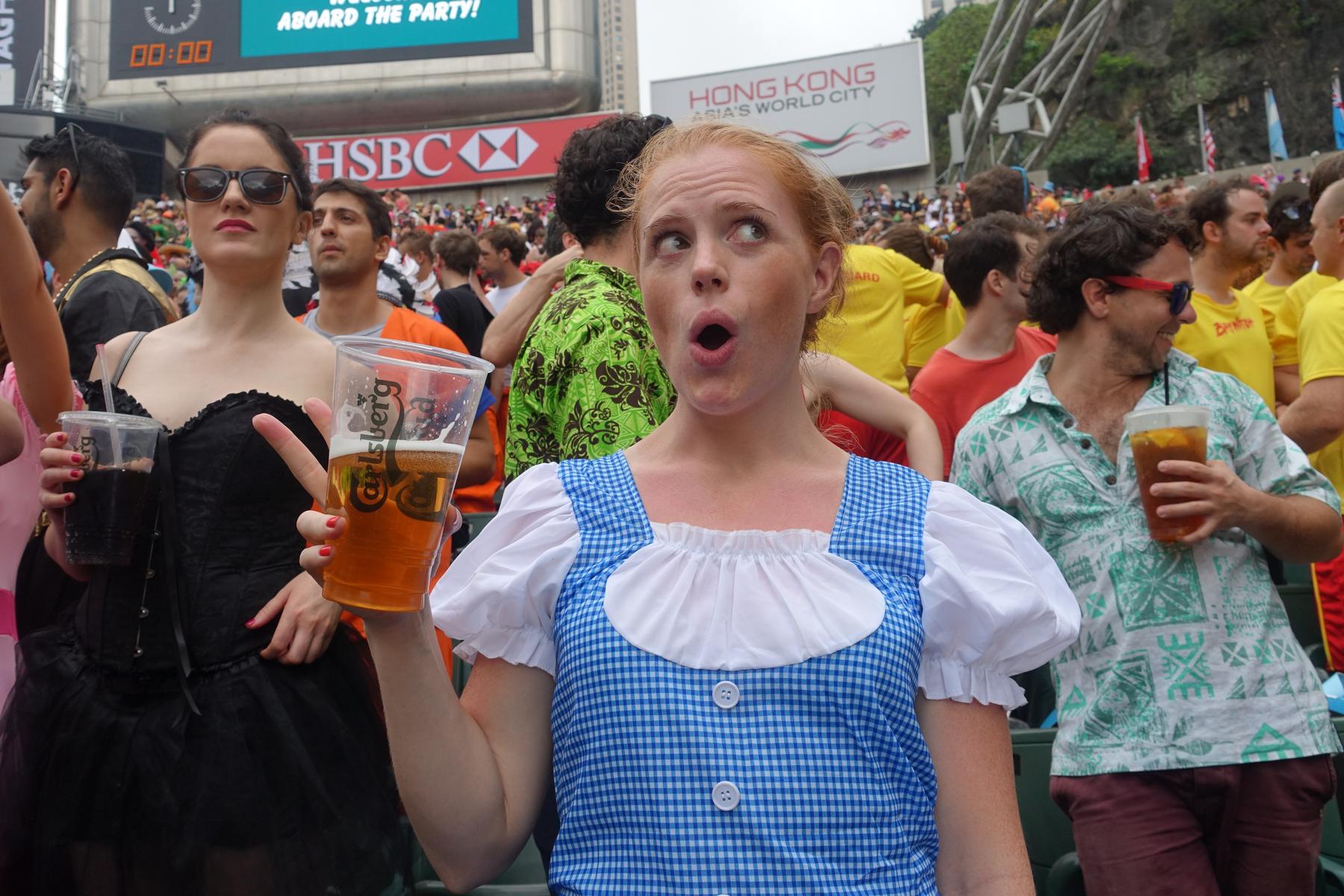 A surprised woman in a Dorothy costume holds a beer among a festive crowd at the Hong Kong Rugby Sevens.