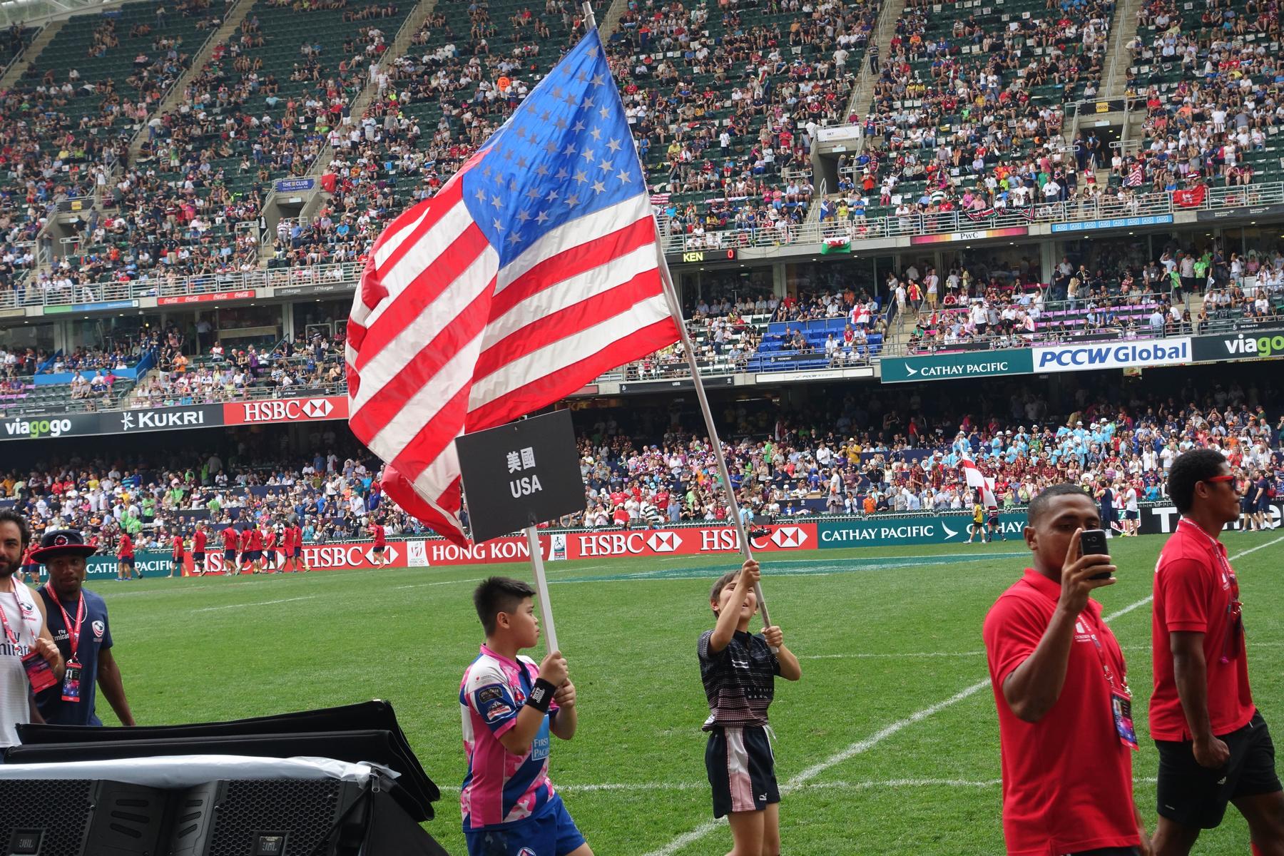 Two children hold an American flag and a "USA" sign on a rugby field packed with spectators at the Hong Kong Sevens.