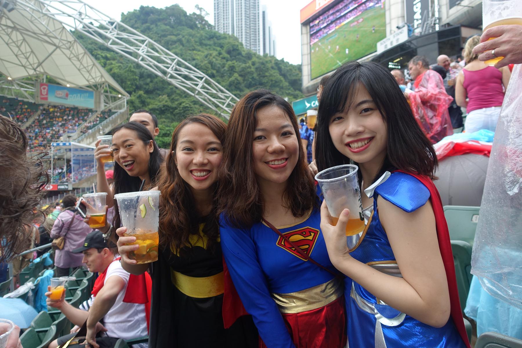 Three smiling women in superhero costumes hold drinks at the Hong Kong Rugby Sevens.