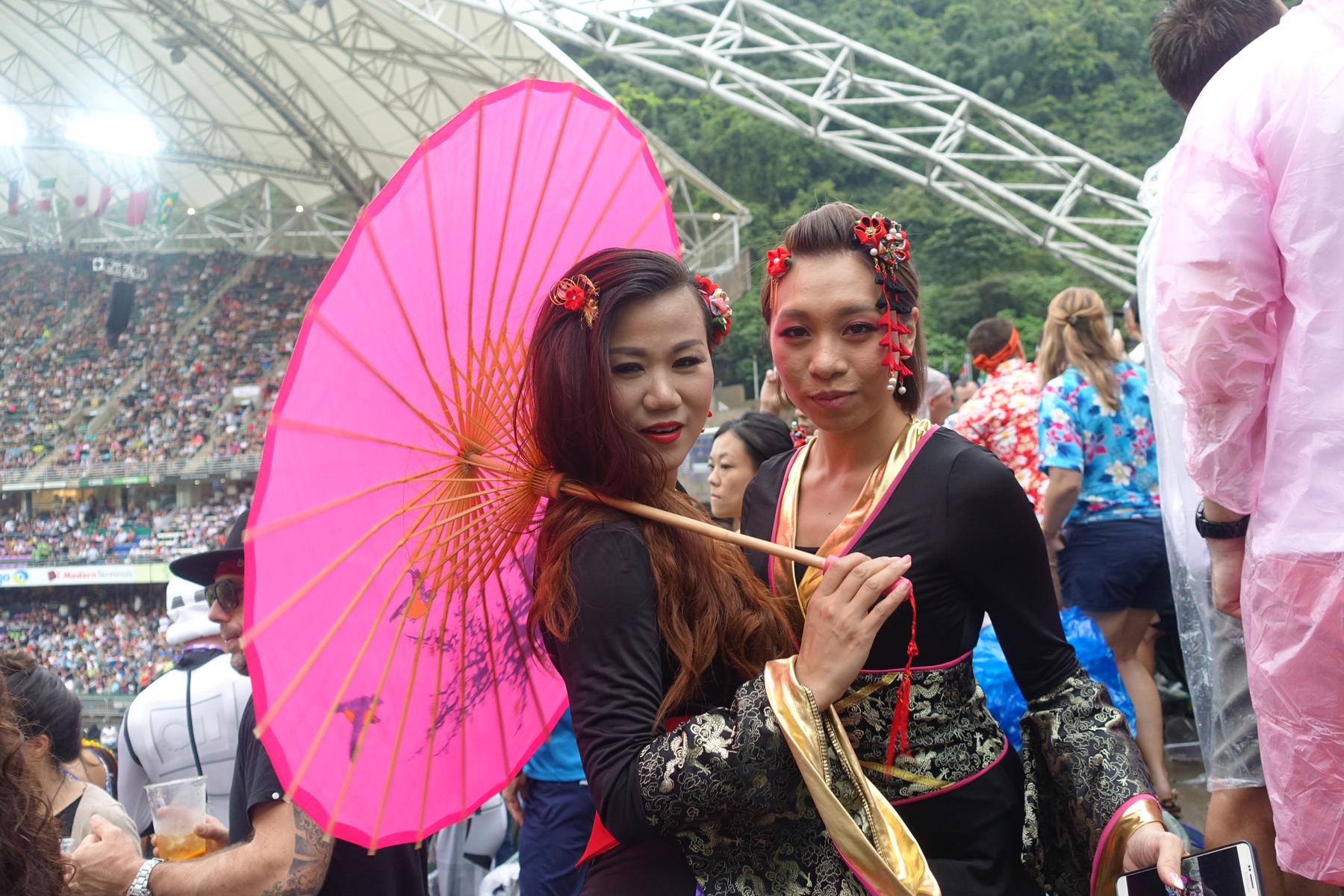 Two women in elaborate traditional costumes, one holding a bright pink parasol, pose at the Hong Kong Rugby Sevens.