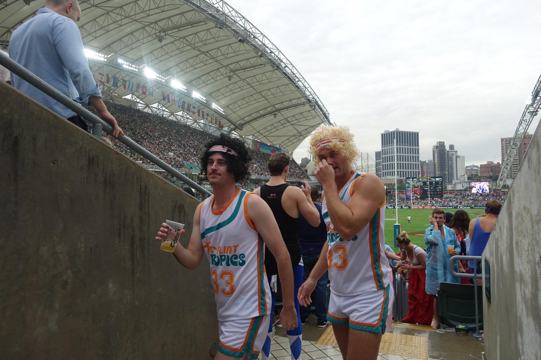 Two fans in "Flint Tropics" costumes and wigs walk through a crowded rugby stadium's South Stand.