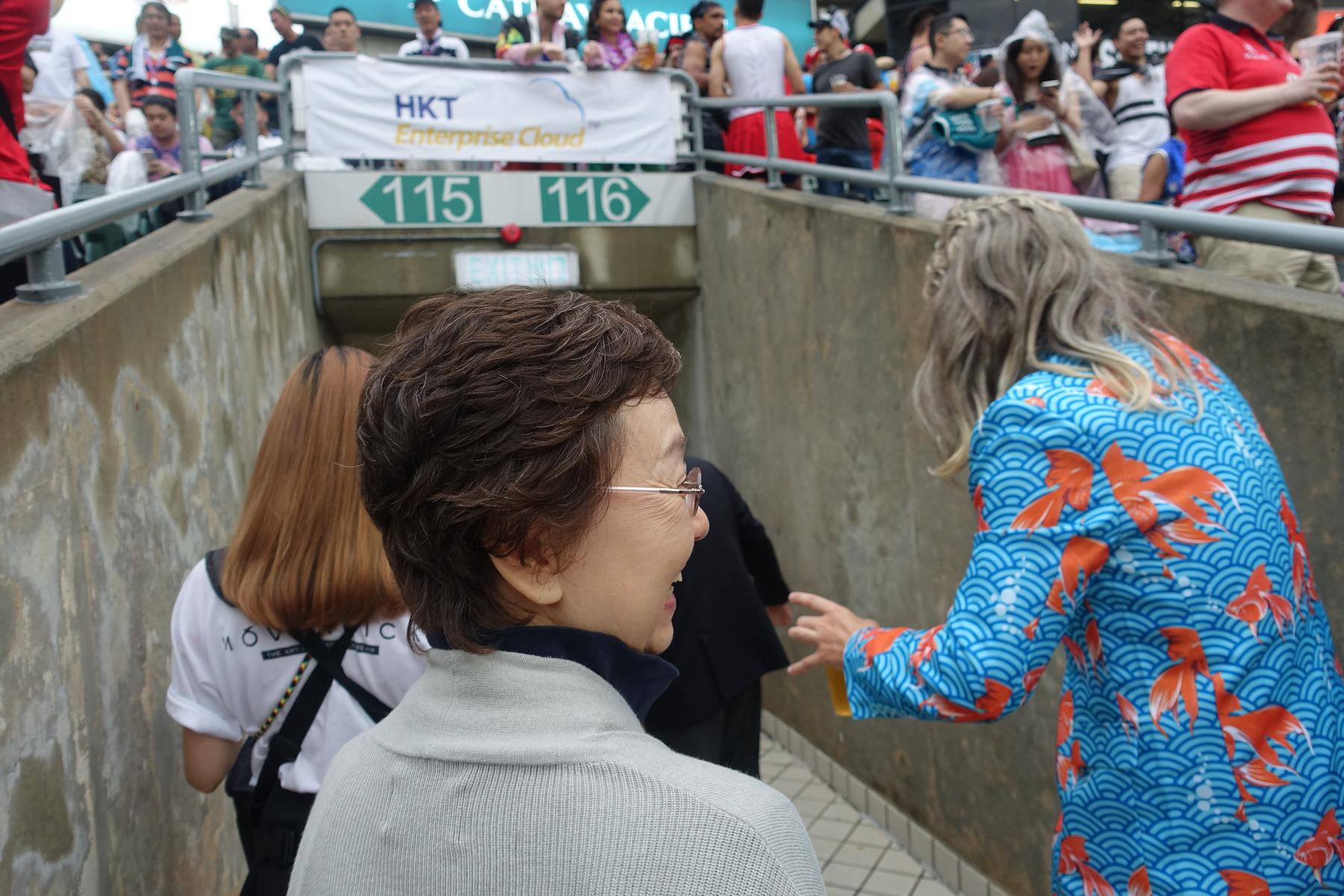 Rugby fans, some in colorful attire like a blue goldfish jacket, move through an entrance to sections 115 and 116 at the Hong Kong Sevens South Stand.