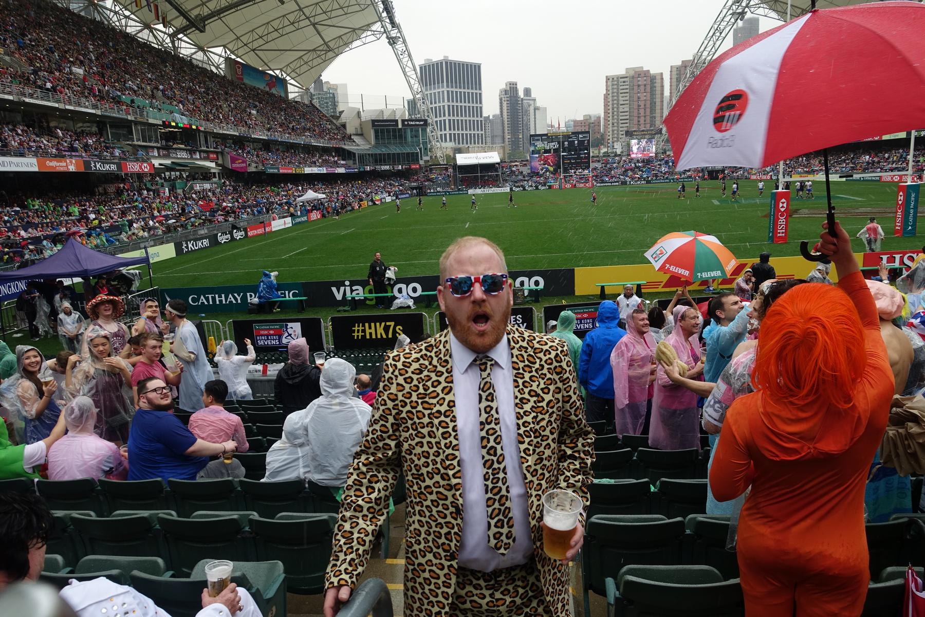 A man in a leopard print suit holds a beer in a crowded, rainy rugby stadium filled with fans in costumes and ponchos.