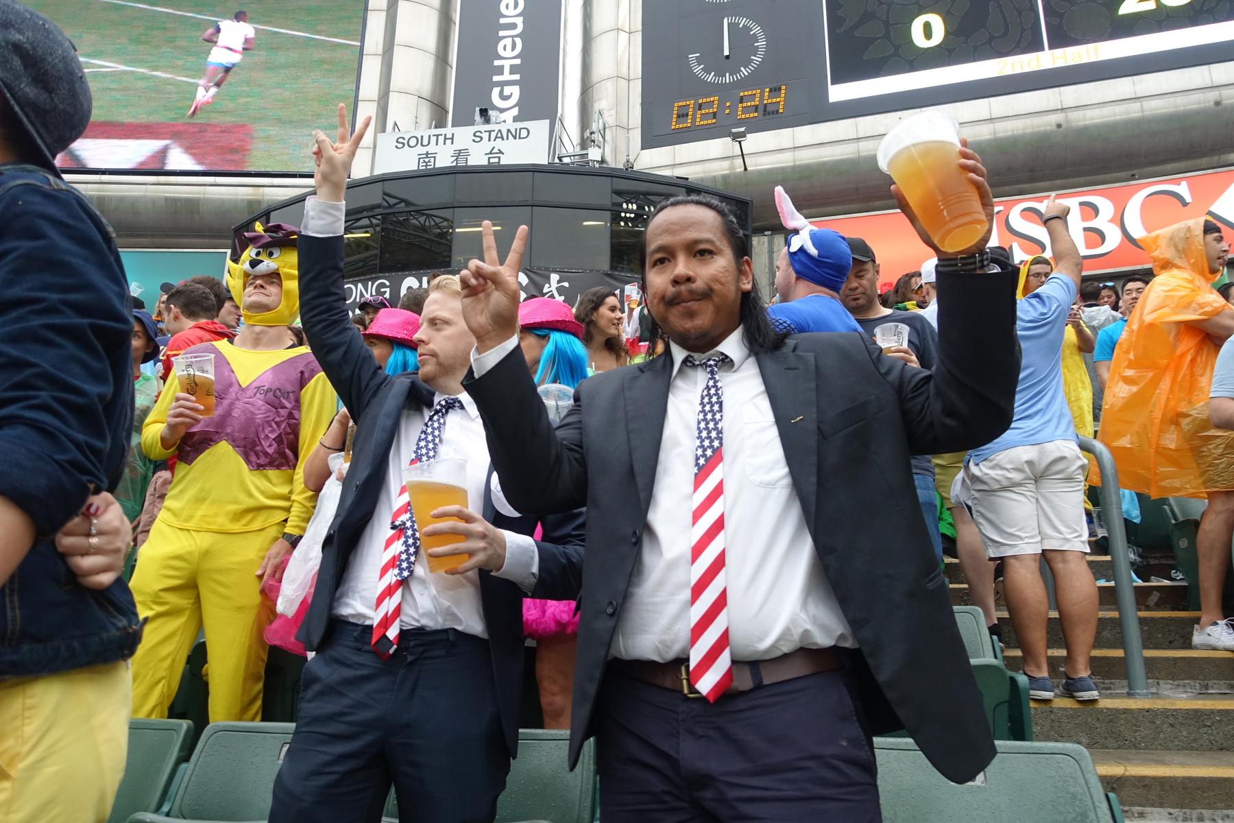 Two men in suits and American flag ties hold beers and make peace signs amidst a costumed crowd at the Hong Kong Rugby Sevens South Stand.