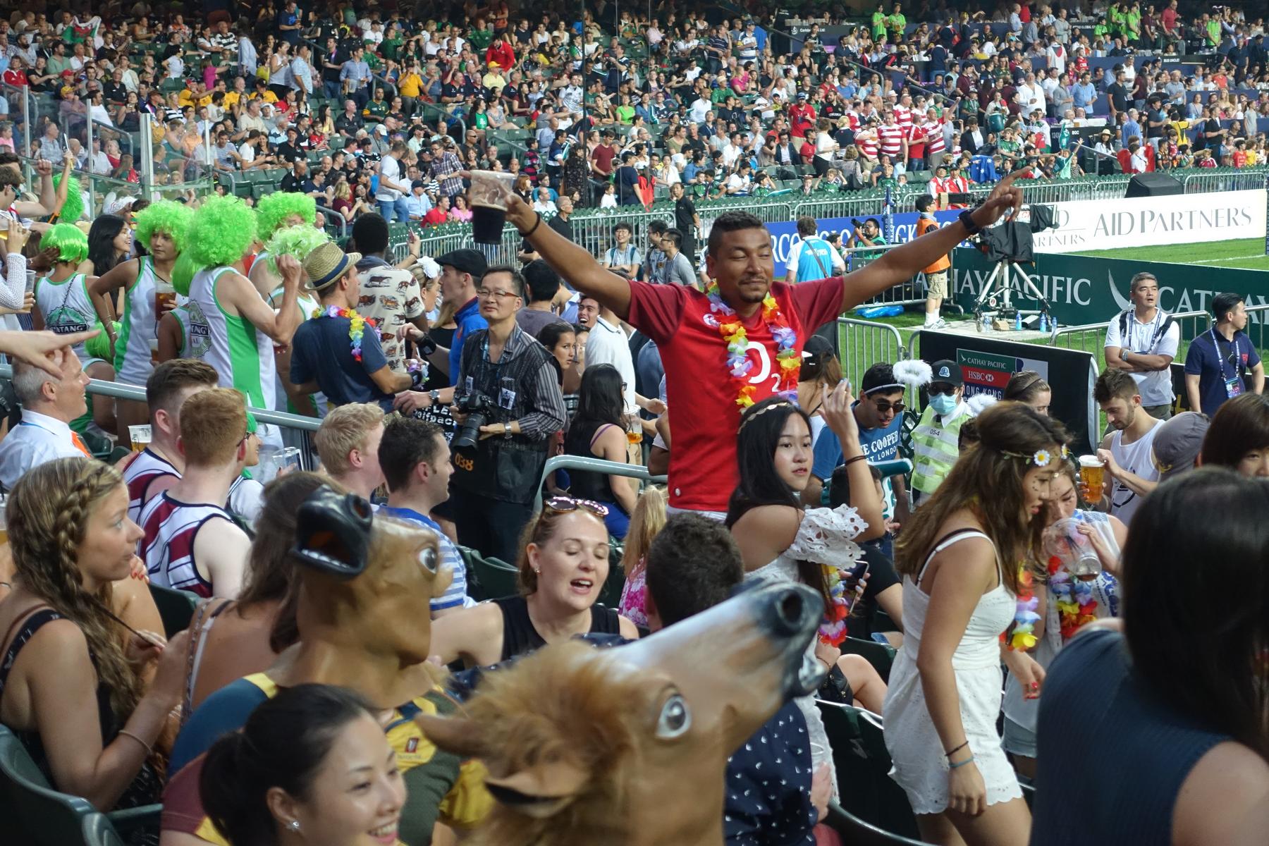 A man in a red shirt and lei stands with arms outstretched in a lively, costumed crowd at the Hong Kong Rugby Sevens.