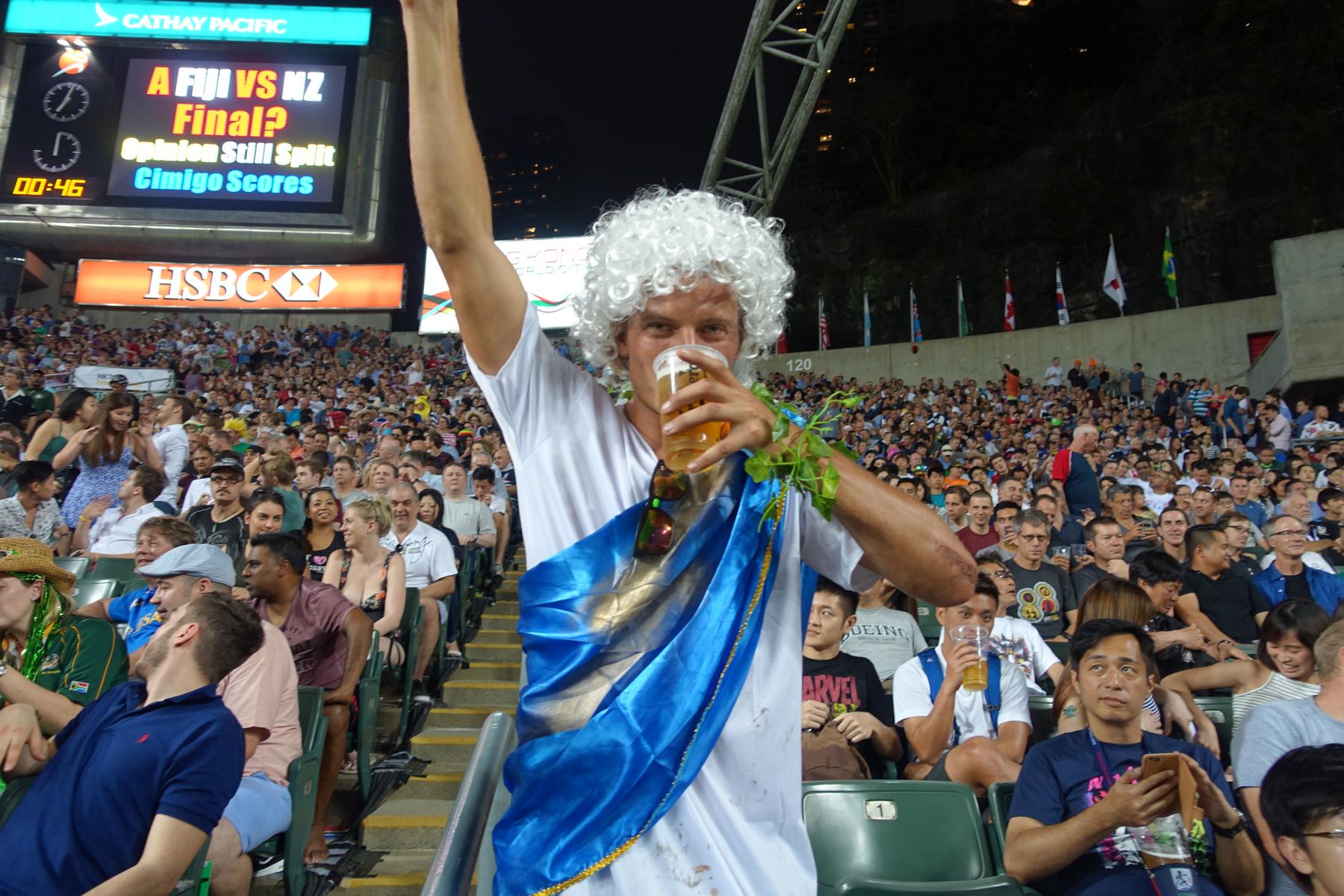 A man in a white wig and blue toga drinks beer and raises an arm in the crowded South Stand of the Hong Kong Rugby Sevens.