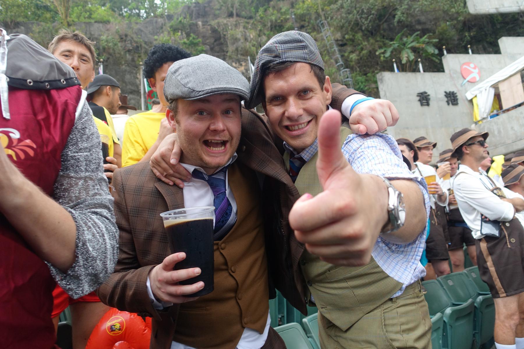 Two cheerful men in flat caps and vests embrace, one holding a dark drink and the other giving a thumbs-up.